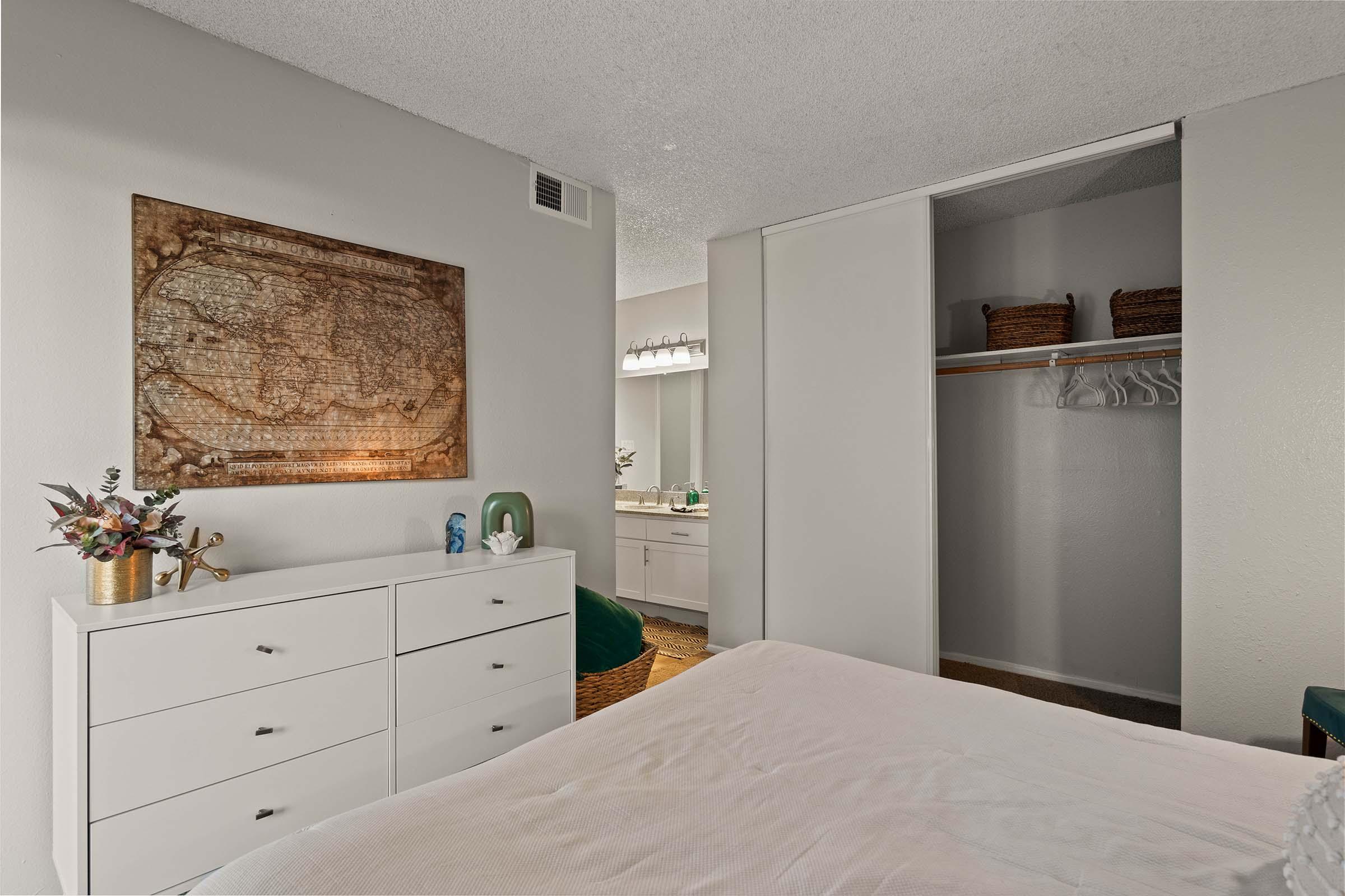 A cozy bedroom featuring a white dresser with decorative items, a world map art piece on the wall, and an open closet with hanging space. In the background, a bathroom area is visible, showcasing a mirror and additional storage. The room has light colors and a welcoming atmosphere.