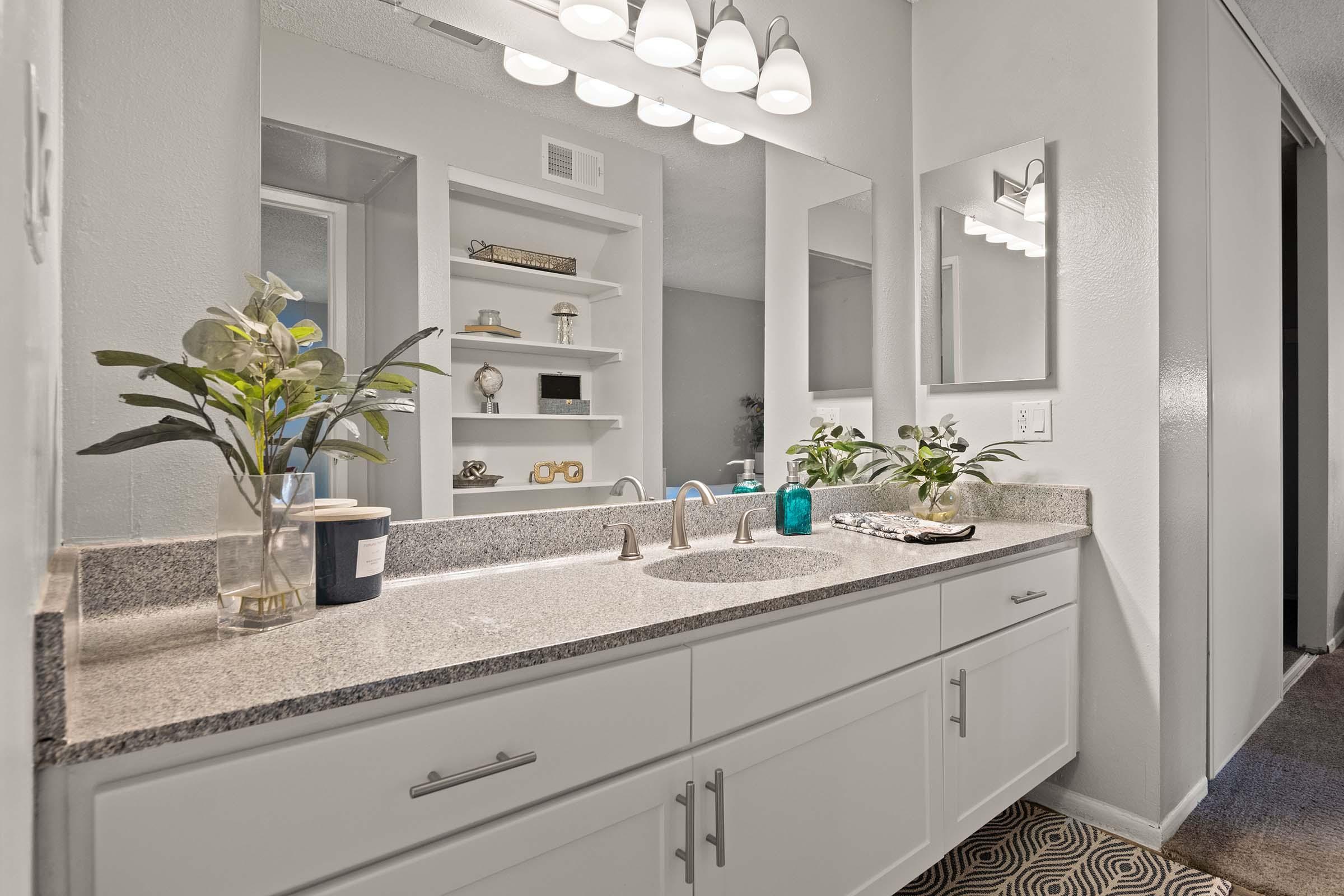 A modern bathroom with a double sink vanity featuring a gray countertop and white cabinets. Above the sinks, there are stylish light fixtures and a wall mirror. On the countertop, there are decorative plants, a blue vase, and toiletries. Shelves in the background display decorative items, adding a touch of elegance.