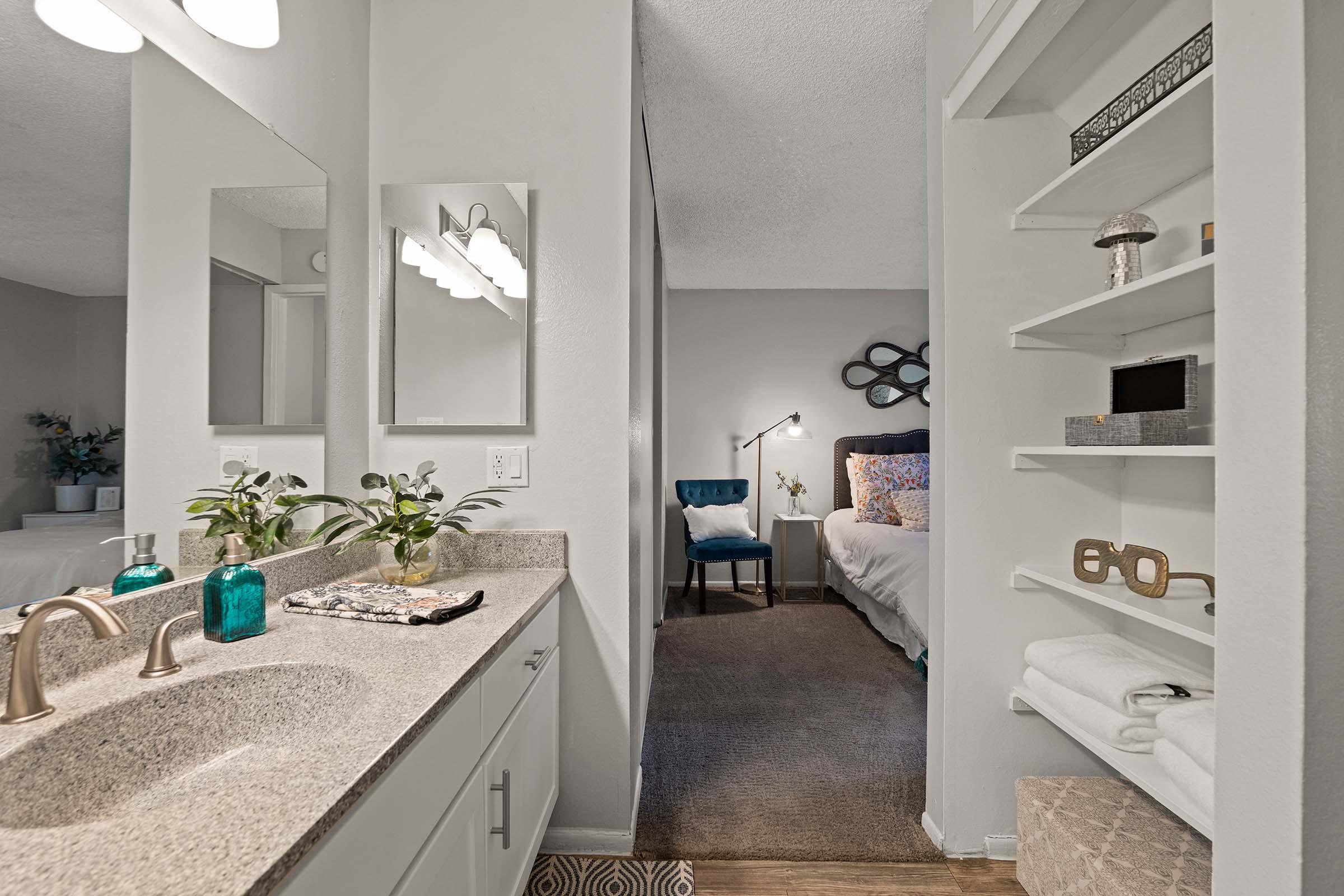 A modern bathroom featuring a double sink with a gray countertop, mirrors above, and a small plant. To the right, a glimpse of a cozy bedroom with a blue chair and decorative elements. White shelves hold towels and decor, and the room is well-lit with soft lighting.