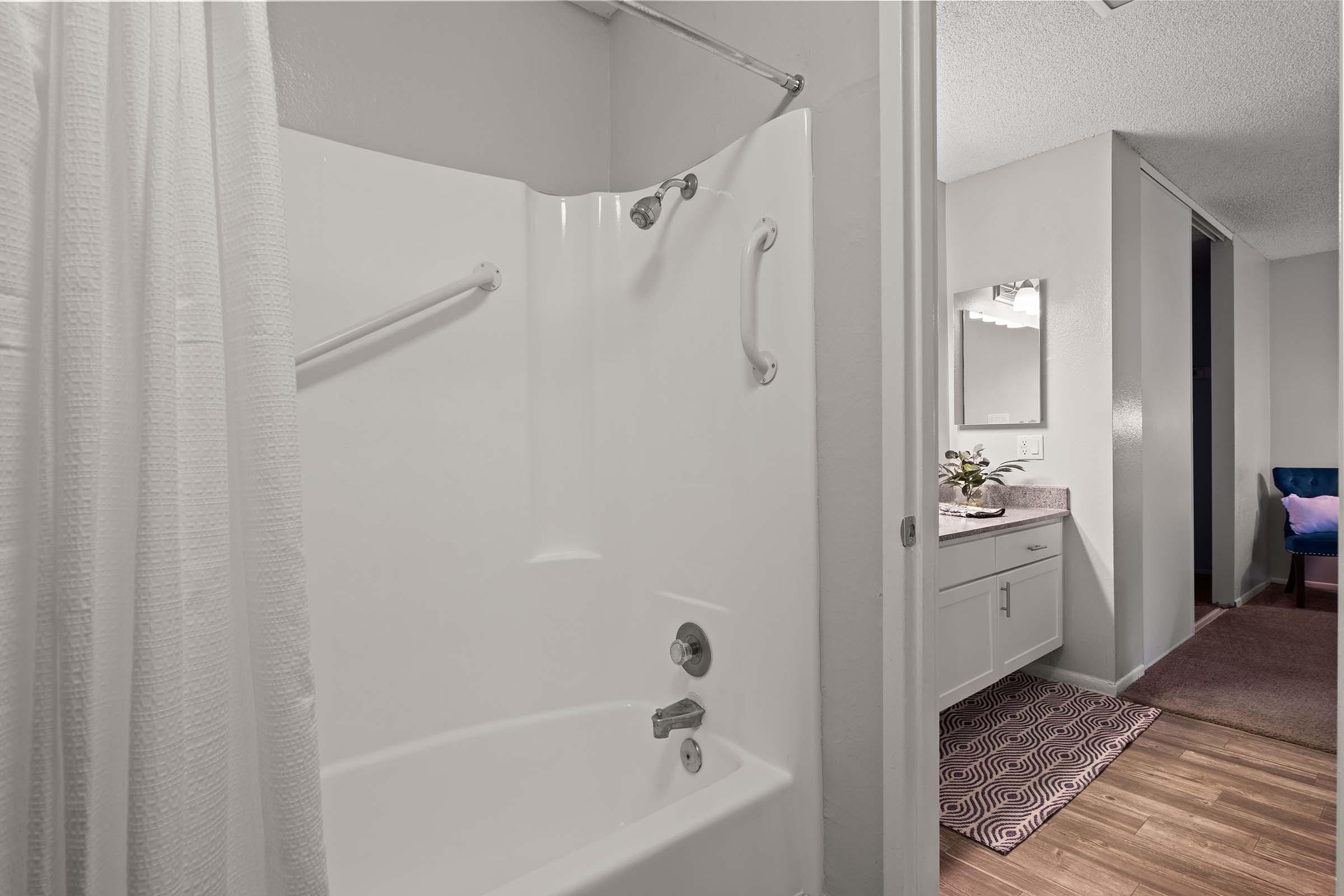 A modern bathroom featuring a white shower stall with a bathtub, grab bars, and a shower curtain. The space includes a vanity with a mirror on the right, and a glimpse of a carpeted area and additional fixtures in the background, creating a clean and functional design.