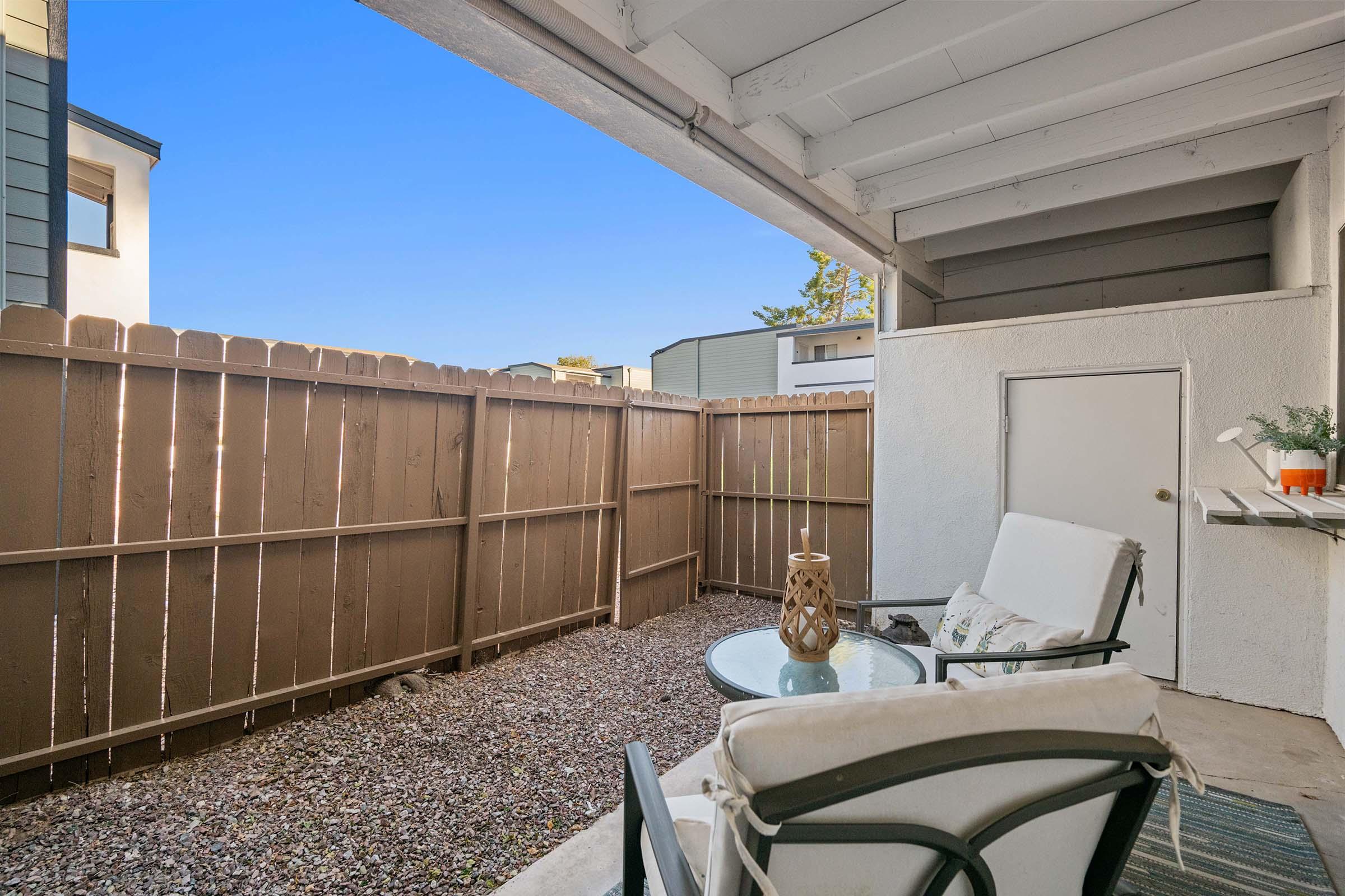 A cozy outdoor patio area featuring two white chairs and a small glass table, surrounded by a wooden fence. There’s a decorative lantern on the table and a door leading to another space. Bright blue sky overhead complements the inviting atmosphere.