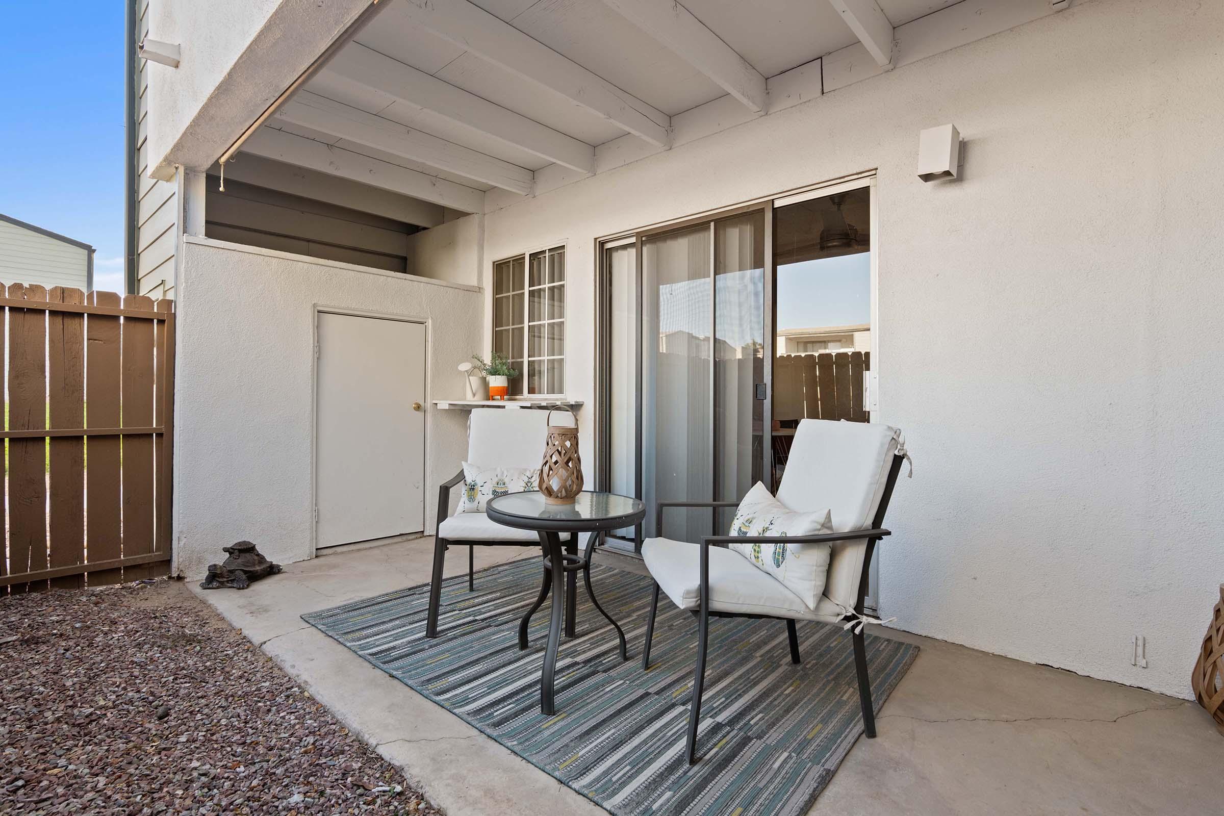 A cozy outdoor patio featuring two white chairs with decorative pillows around a small black table, situated on a gravel floor. There is a sliding glass door leading inside, and a wooden fence in the background, creating a private and inviting space for relaxation.
