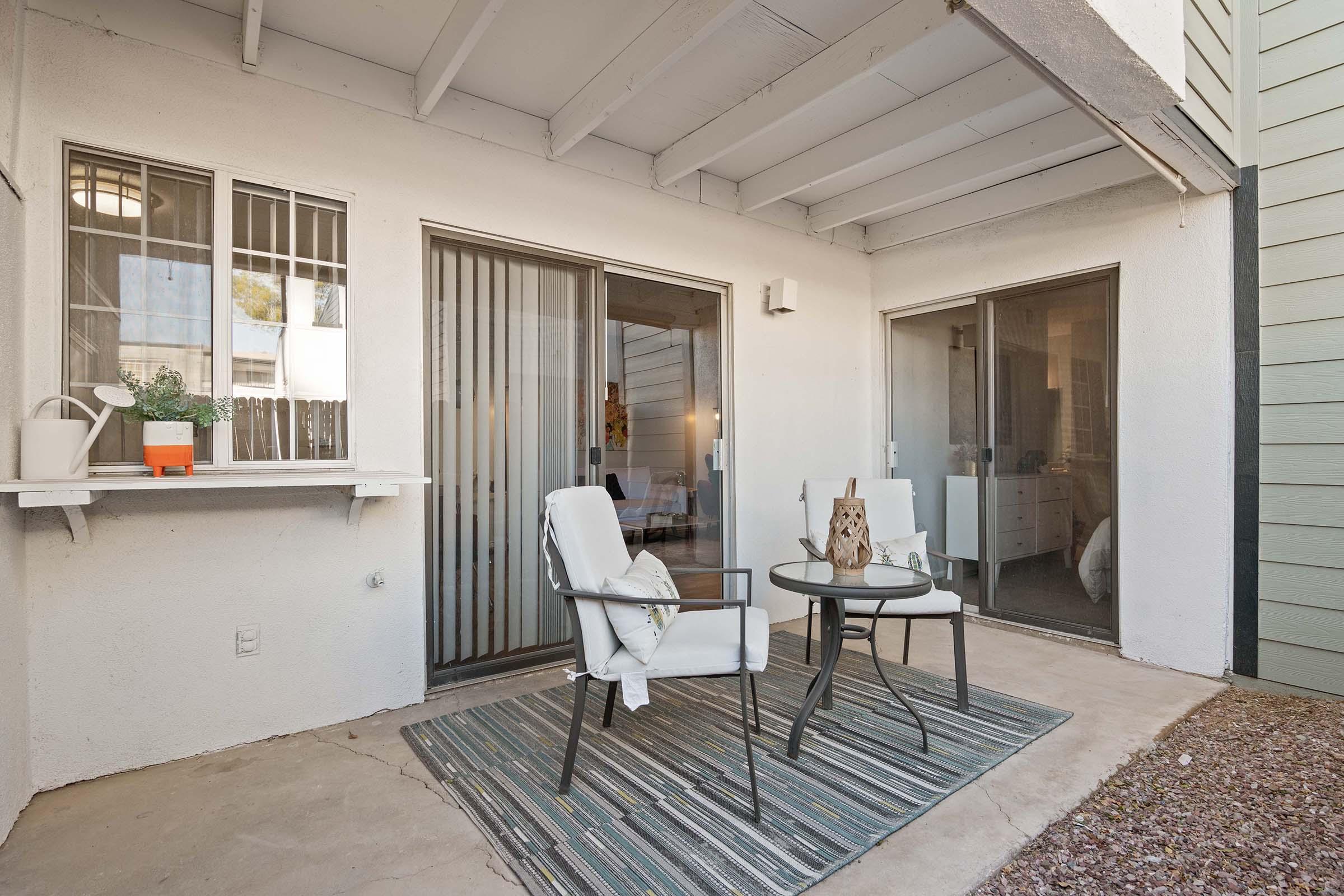 A cozy outdoor patio area featuring two white chairs and a small black table on a striped rug. The space is complemented by a potted plant on a shelf and has glass sliding doors leading to an indoor living area, enhancing the inviting atmosphere. The walls are neutral-toned with a rustic ceiling.