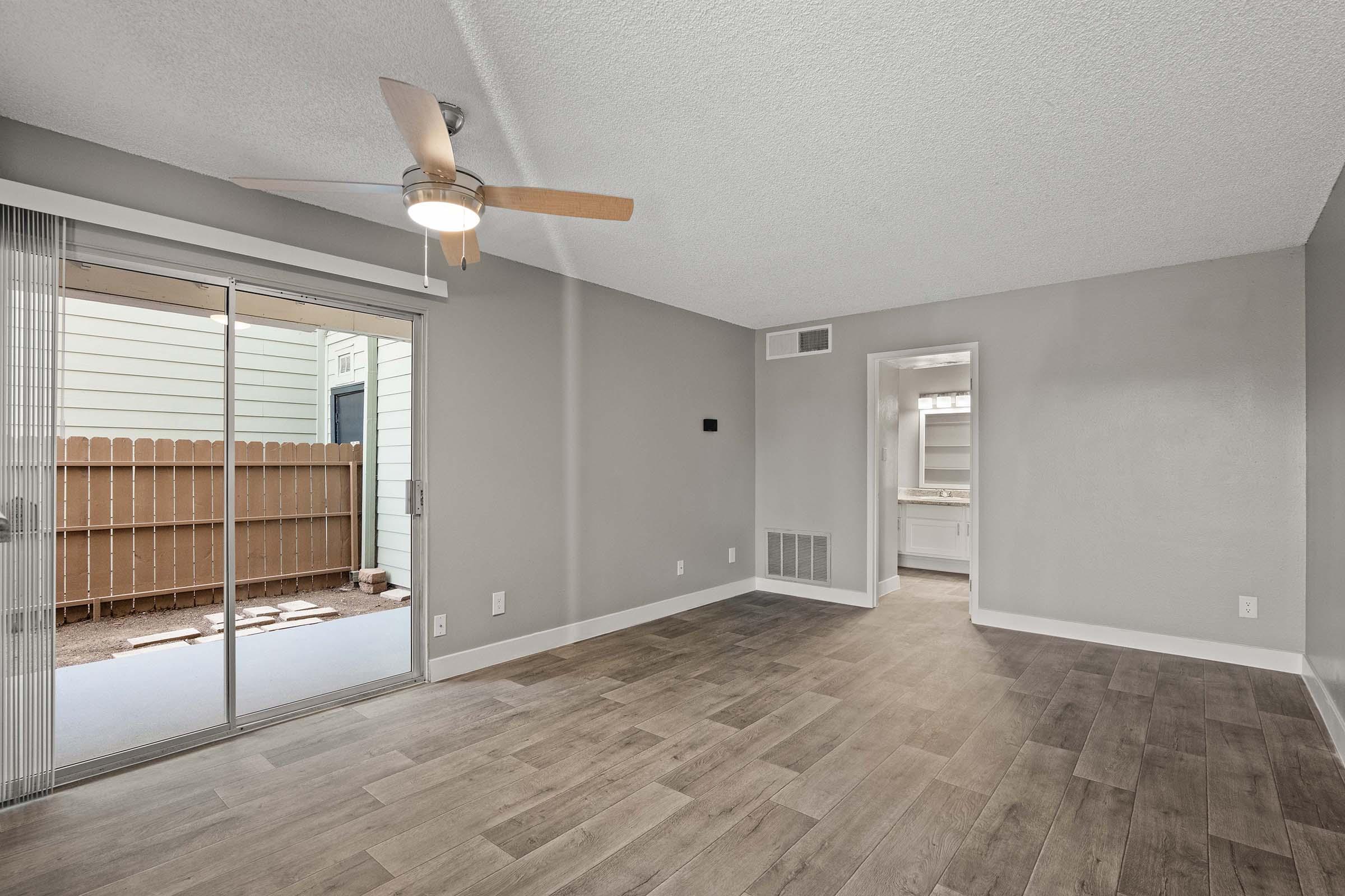 A spacious living room with a ceiling fan, neutral gray walls, and light wood flooring. There is a sliding glass door leading to a small outdoor area with a fenced yard. A doorway on the right leads to a kitchen area. Natural light fills the room, creating a bright and open atmosphere.