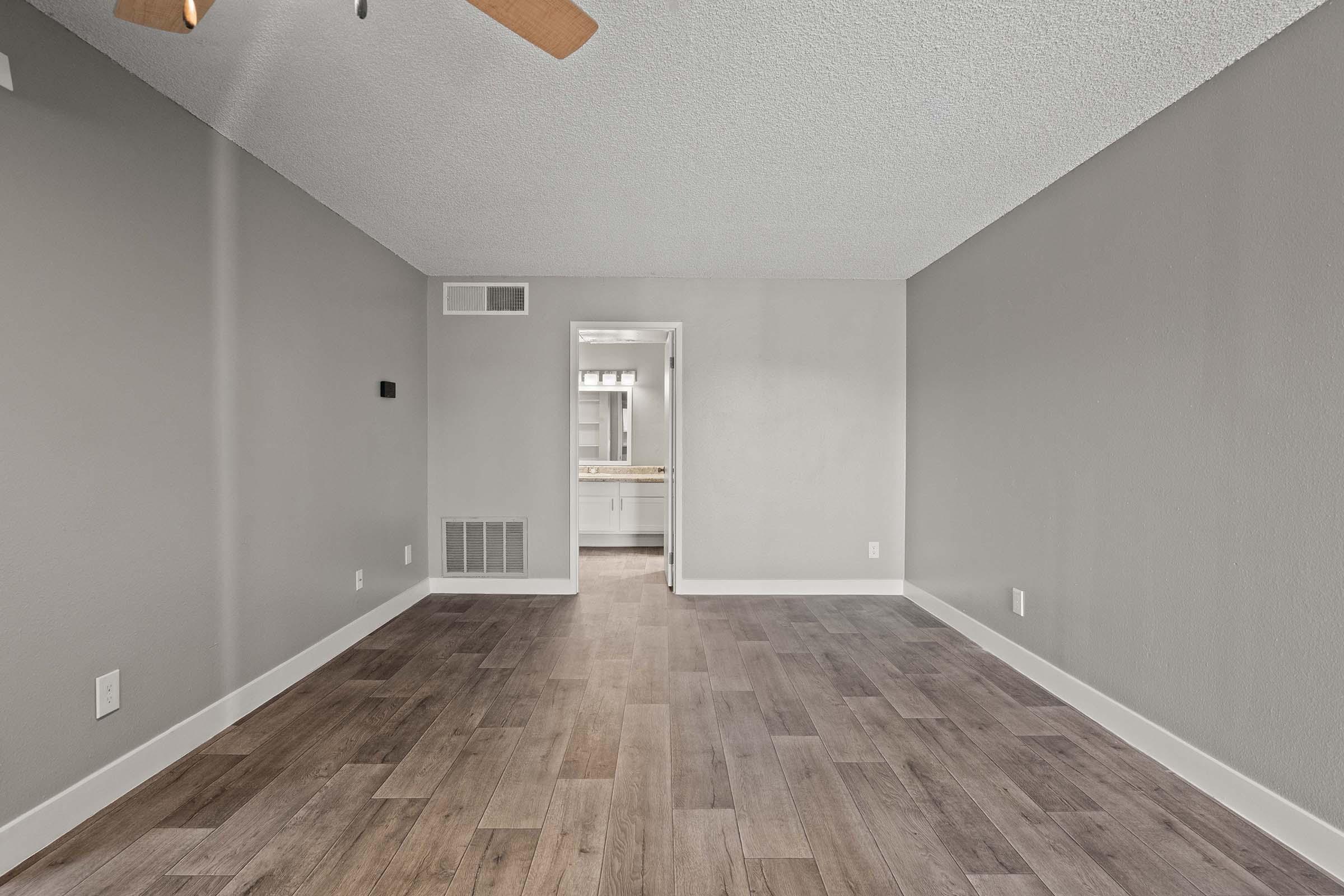 A spacious, empty room with light gray walls and wooden flooring. A ceiling fan is visible, and there is an open doorway leading to a bathroom in the background. The room has a minimalist appearance with no furniture or decorations. Natural light may be entering from a window outside the frame.