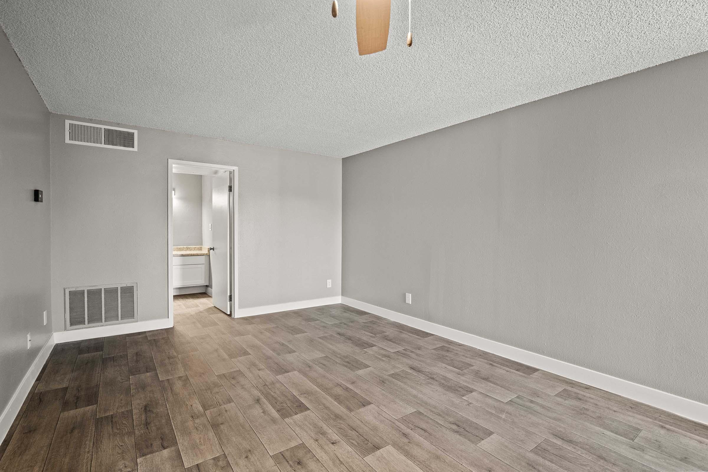 A spacious room with a light gray textured ceiling and neutral-colored walls. The floor is covered in wooden planks. A ceiling fan hangs from the ceiling, and a doorway leads to a bathroom area in the background, suggesting an inviting and modern living space.