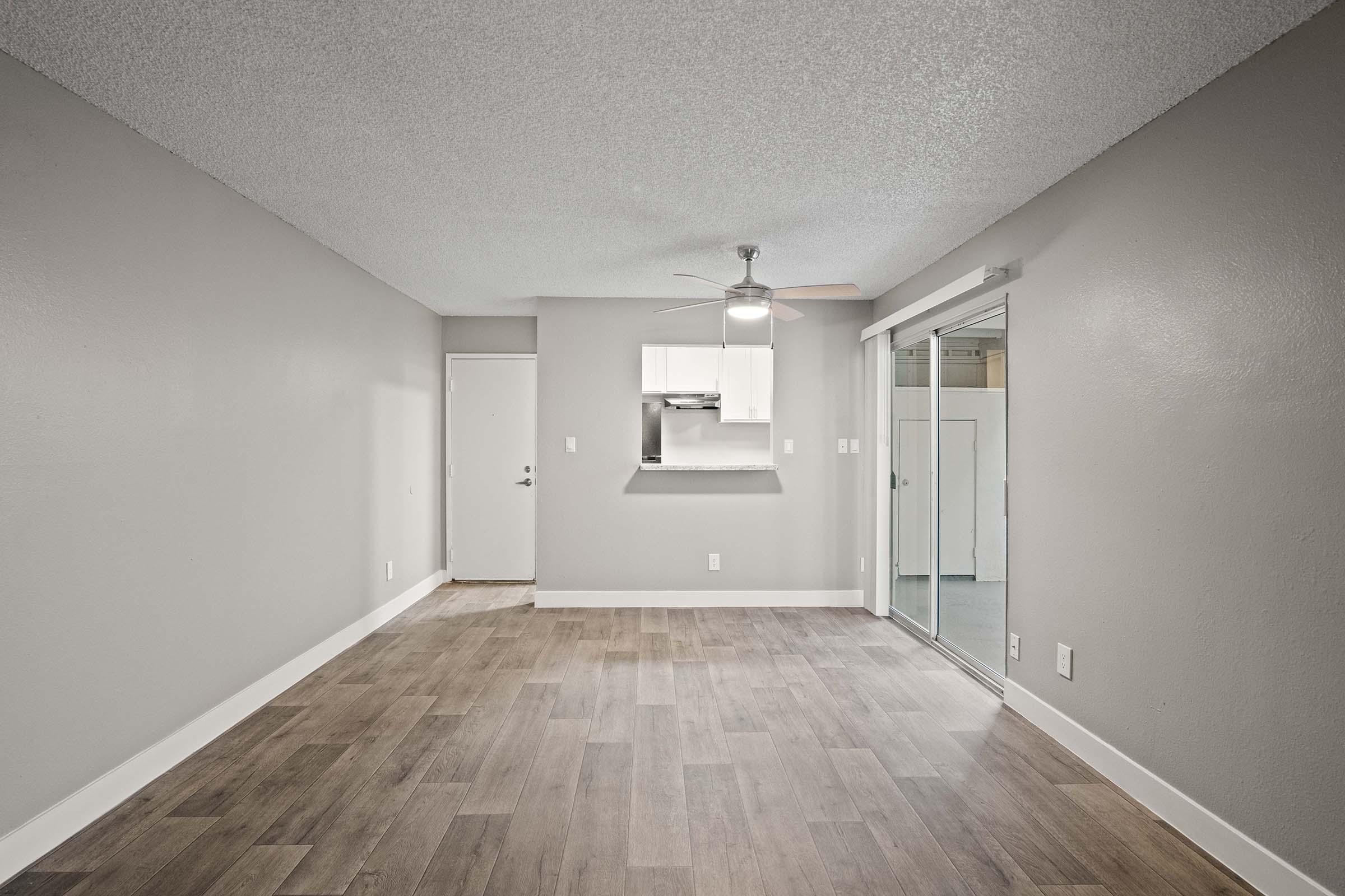A spacious, empty living room featuring light gray walls and a wood-look floor. A ceiling fan is visible, and a sliding glass door leads to an outdoor area. The kitchen area can be seen in the background, separated by a counter. The room has a clean and modern appearance, inviting natural light.