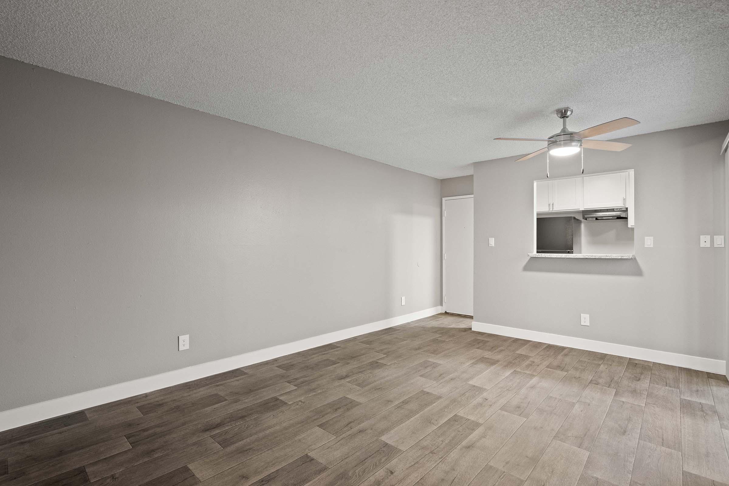 A spacious, empty room with light gray walls and a textured ceiling. The floor features wooden planks, and there's a ceiling fan with light. An open kitchen area is visible in the background, separated by a short breakfast bar. A door leads to another room on the left side of the image.