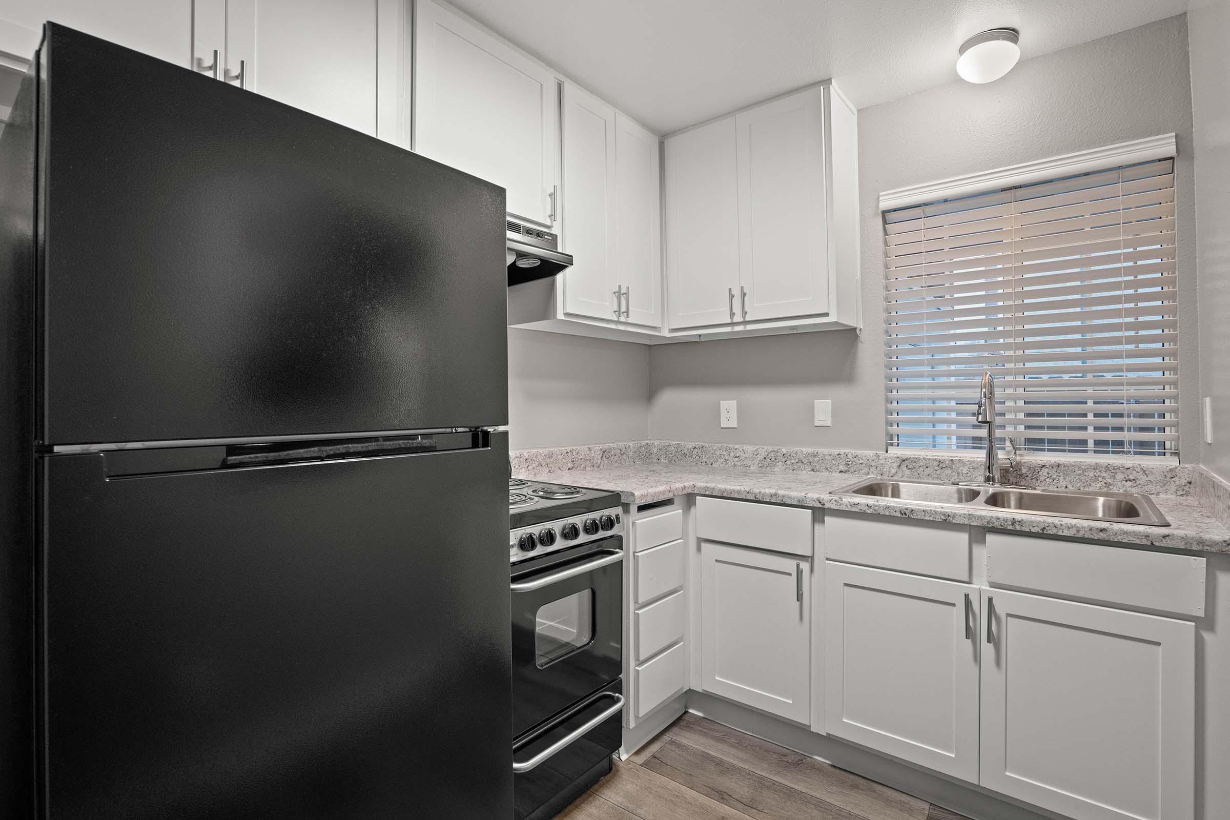 A modern kitchen featuring a black refrigerator and an oven with a gas stove. The cabinets are white, and the countertops are light-colored granite. A window with blinds allows natural light, and there are dual sinks under the window. The flooring is wooden, enhancing the contemporary design of the space.