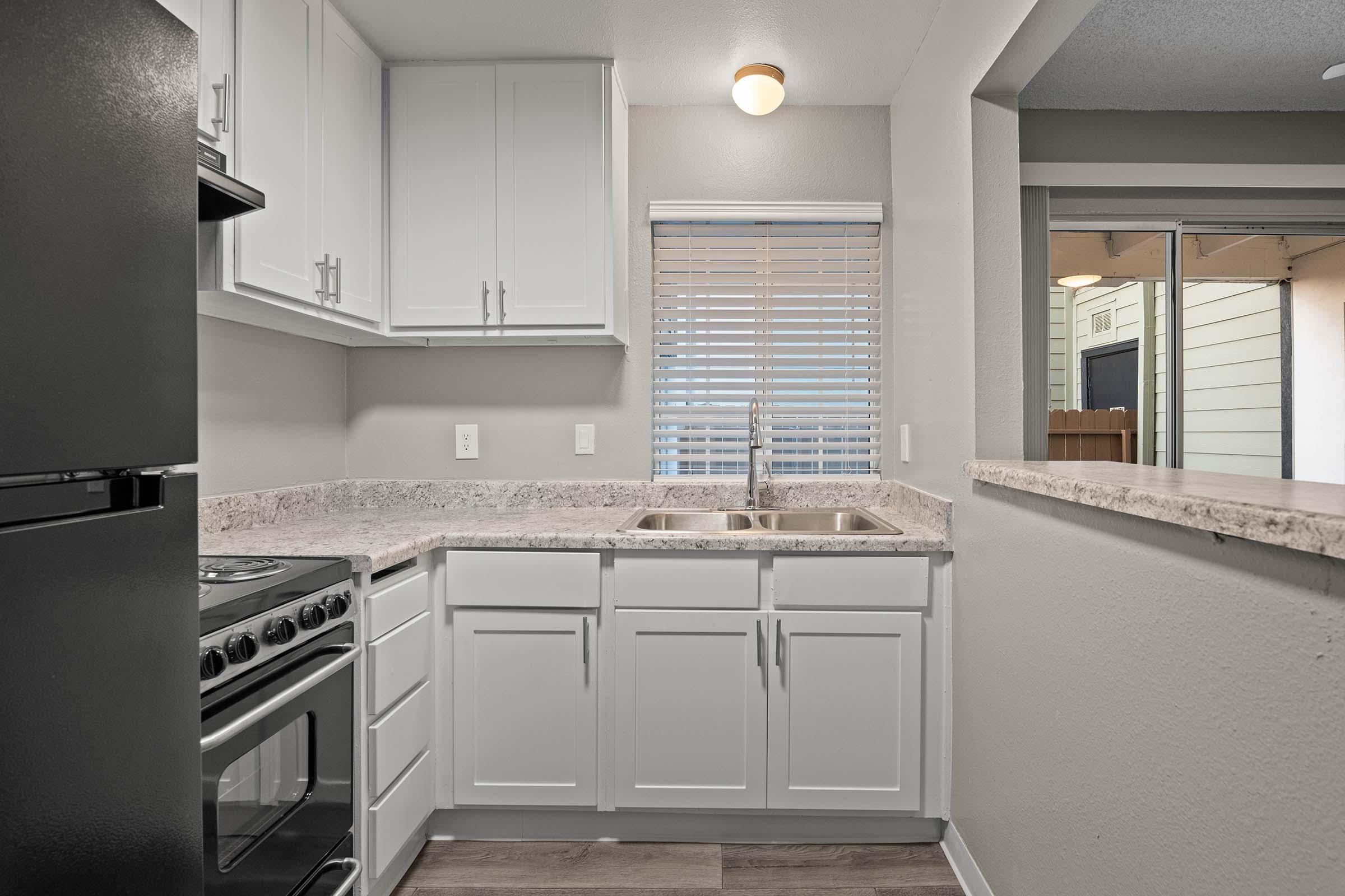 Modern kitchen featuring white cabinetry, a granite countertop, a black stove, and a double sink under a window with blinds. The space is well-lit with overhead lighting and has an open layout connecting to a living area. The flooring is a light wood laminate.