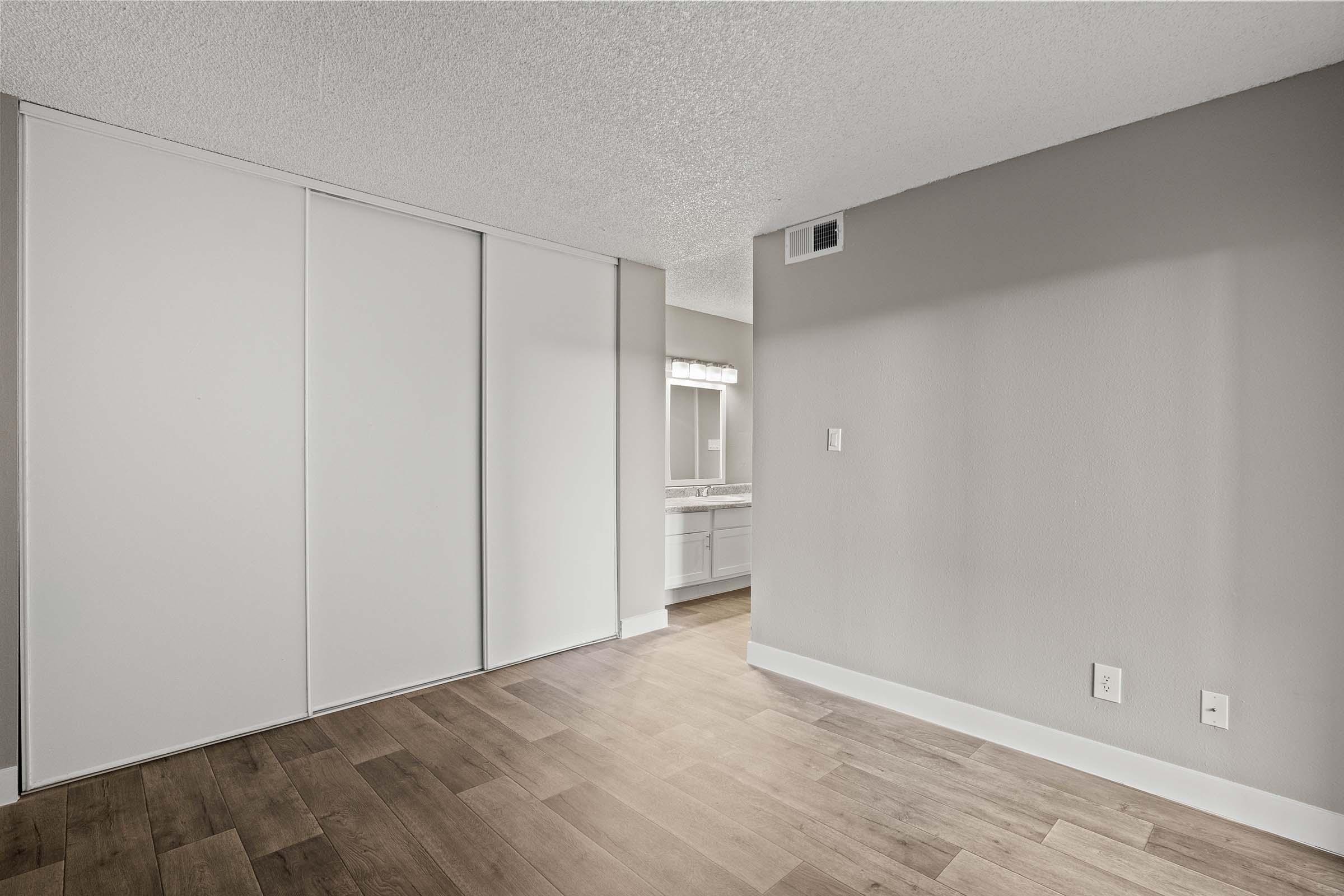 A well-lit interior space featuring light-colored walls and wooden flooring. On the left, there is a closet with sliding doors, and on the right, a bathroom area with a vanity mirror and sink visible in the background. The room has a modern and clean aesthetic, with ample natural light.