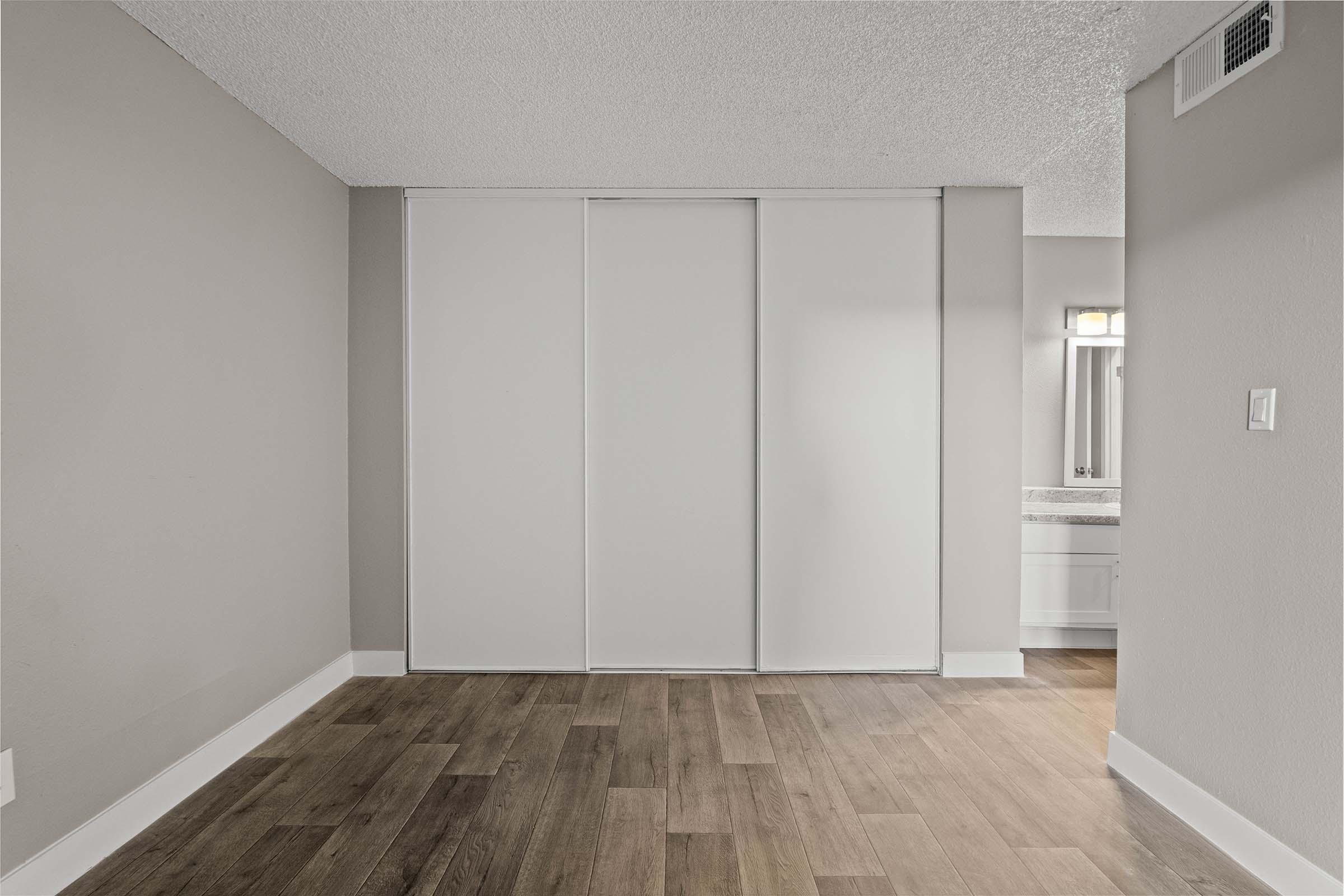 Interior view of a room featuring a light gray wall and wooden flooring. A large closet with sliding white doors is on the left, and a bathroom area with a mirror and vanity is visible in the background. The room appears bright and spacious, with soft lighting.