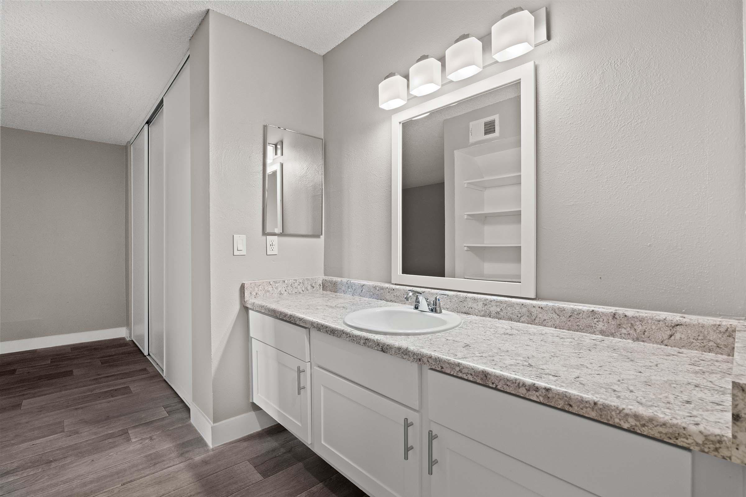 A modern bathroom featuring a white vanity with a granite countertop and a sink, framed mirror above, and three wall-mounted light fixtures. The walls are painted in a neutral tone, and there is a sliding door closet on the left. The floor is finished with dark wood laminate.