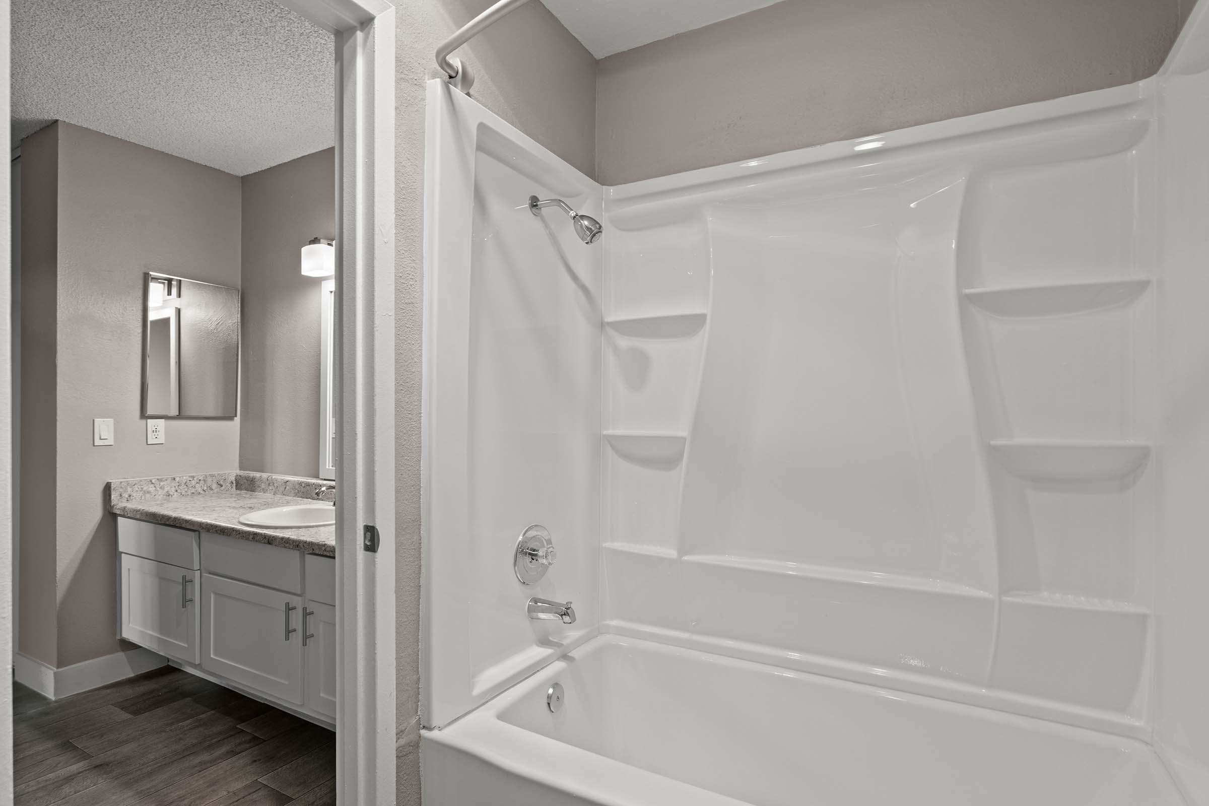 Modern bathroom featuring a clean white bathtub and shower combo, with a granite countertop vanity in the background. A mirror is mounted above the sink, and the walls are painted in neutral tones, creating a bright and spacious atmosphere. The flooring is dark, adding contrast to the light fixtures and surfaces.