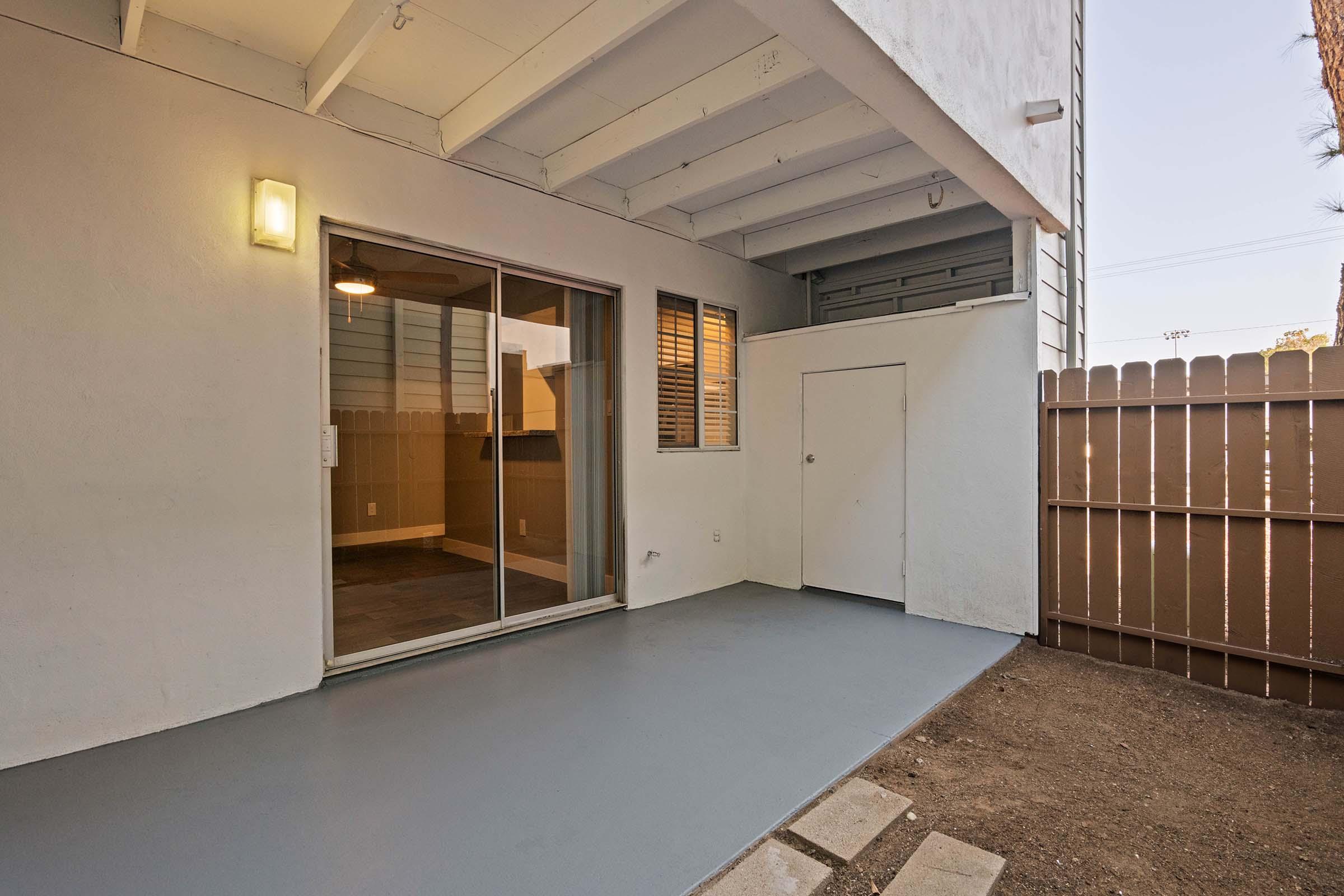A patio area with a gray floor, sliding glass doors leading into an interior space, and a small fenced yard. The walls are white, and there is a light fixture on the wall. The area appears clean and well-maintained, with natural light coming in through the doors.