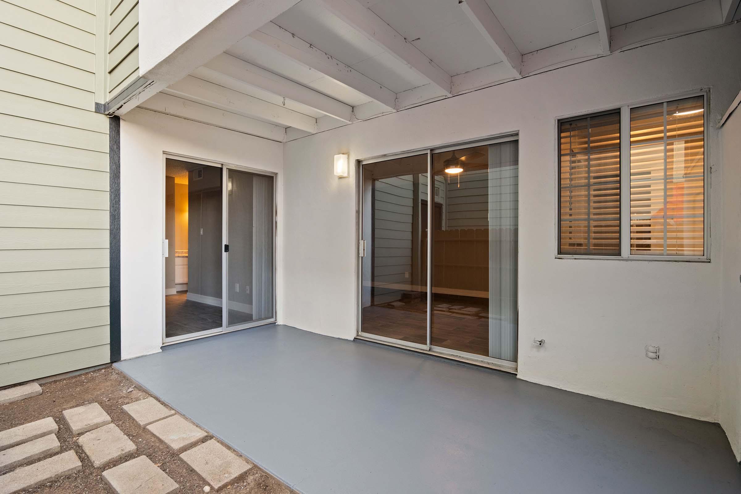 Interior patio area with a smooth gray floor, featuring sliding glass doors on two sides. One side opens to an interior space, while the other leads to an outdoor area. The walls are painted white, and there's a ceiling with exposed beams. Natural light enters from the doors and windows.