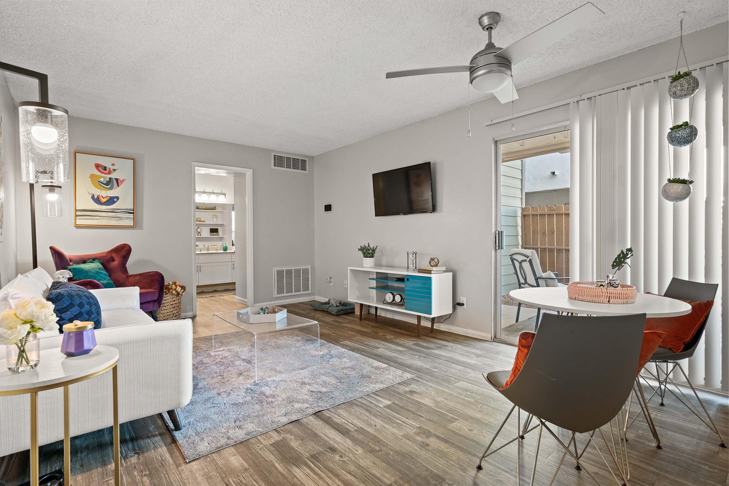 Cozy living room featuring a white sofa with colorful throw pillows, a small round dining table with modern chairs, and a wall-mounted TV. Natural light streams through vertical blinds, illuminating a light grey rug on a brown wood floor. A sliding door leads to an outdoor area, adding to the inviting atmosphere.