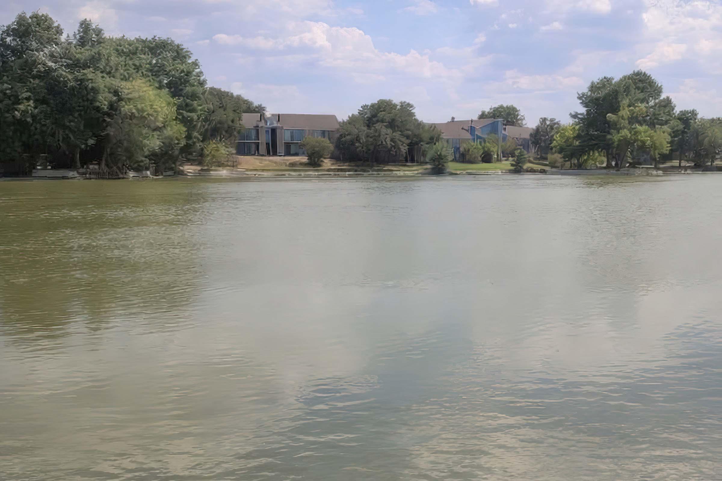 A calm lake reflecting the sky, surrounded by trees and residential buildings. The water is still, with a few ripples, and the area appears peaceful, with scattered clouds above.