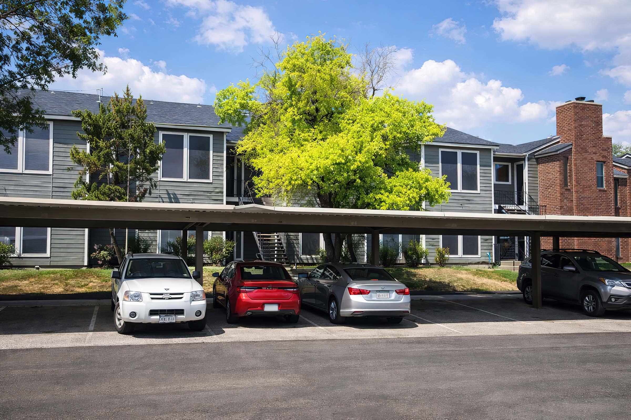 A row of parked cars under a covered area in front of a two-story apartment building. The building features gray siding, large windows, and a staircase leading to the upper level. A vibrant green tree provides shade, and the sky is partly cloudy.