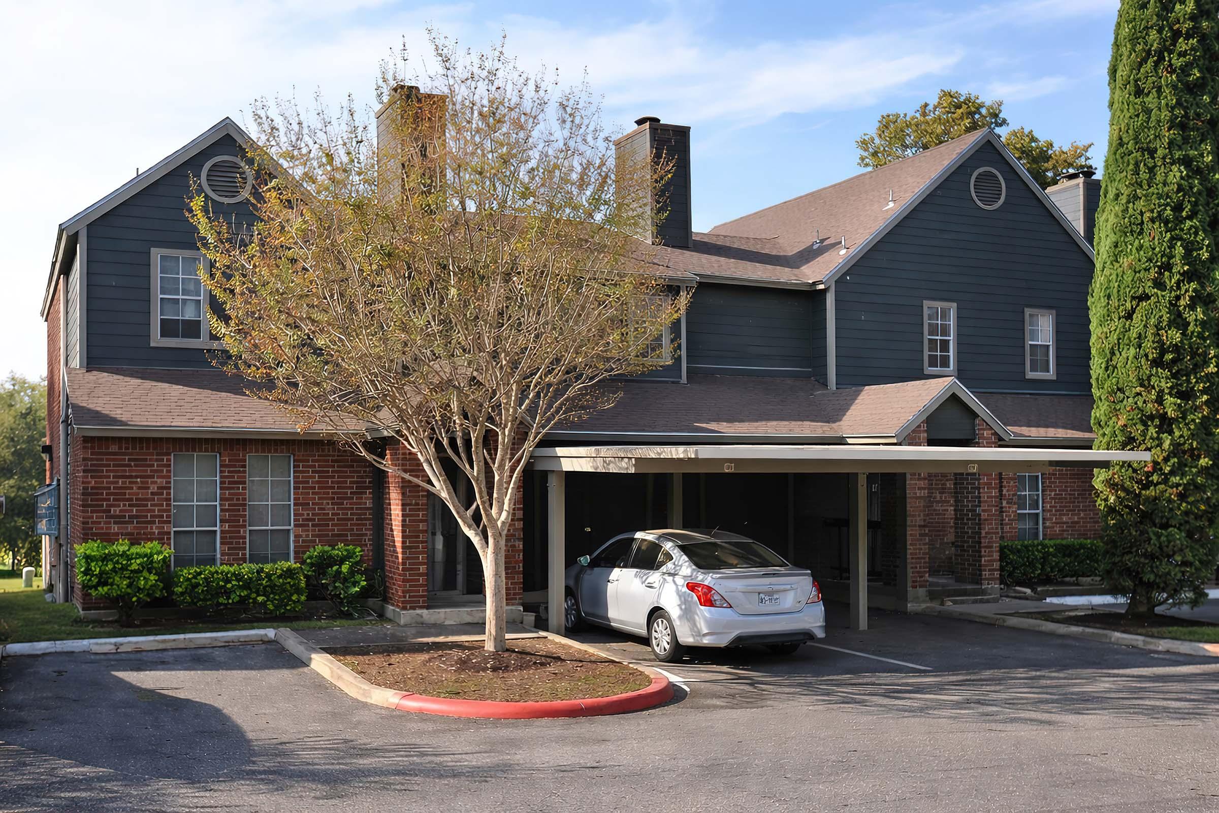 A two-story building featuring a mix of brick and blue siding, with a tree in the front yard and a parked silver car beneath a carport. The scene is set on a clear day, with green bushes lining the walkways, providing a residential atmosphere.