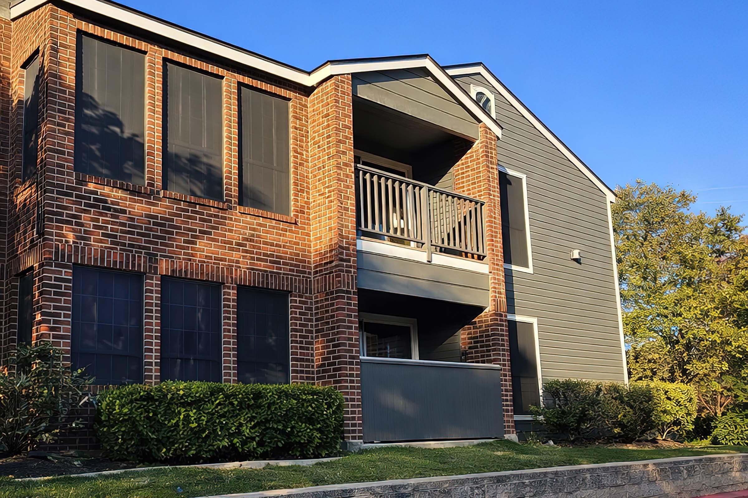 A three-story residential building featuring brick exterior on the lower level and gray siding on the upper level. It has balconies with wooden railings, several windows with screens, and landscaped bushes in front. The scene is set against a clear blue sky.