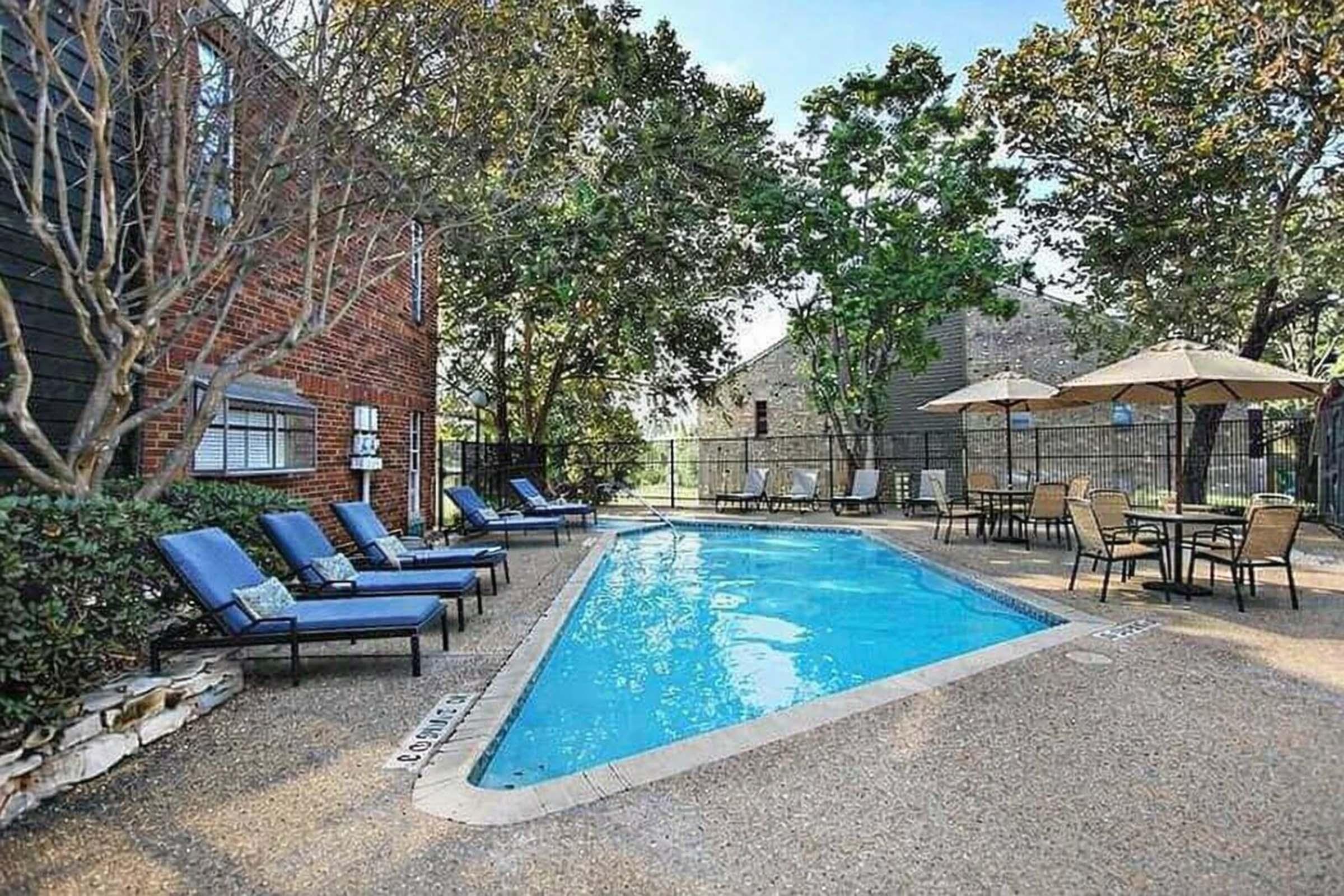 A serene outdoor swimming pool area surrounded by lounge chairs and umbrellas. Lush greenery and trees provide shade, while a cozy seating arrangement is visible nearby. The pool is clear and inviting, offering a relaxing atmosphere in a residential setting.