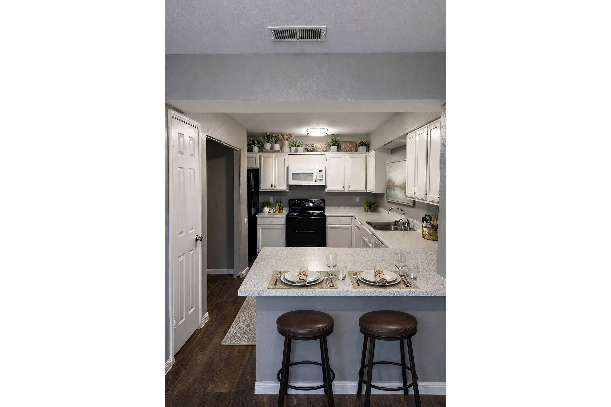 A modern kitchen featuring white cabinetry, black appliances, and a light gray countertop. Two wooden stools are positioned at a kitchen island, set with elegant place settings. The space is well-lit, with decorative plants on shelves and a cozy atmosphere. A doorway leads to another room.