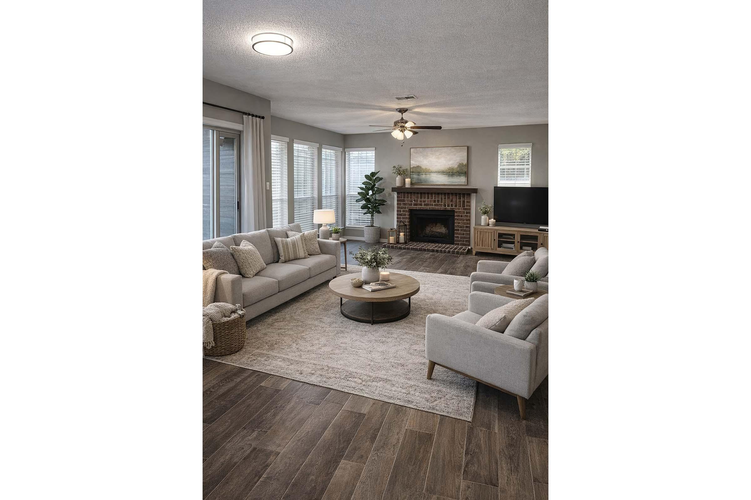 A modern living room featuring light gray sofas and armchairs, a round coffee table, and a cozy fireplace. Large windows let in natural light, and a green plant adds a touch of color. The space is decorated with a neutral area rug, wooden flooring, and soft lighting.