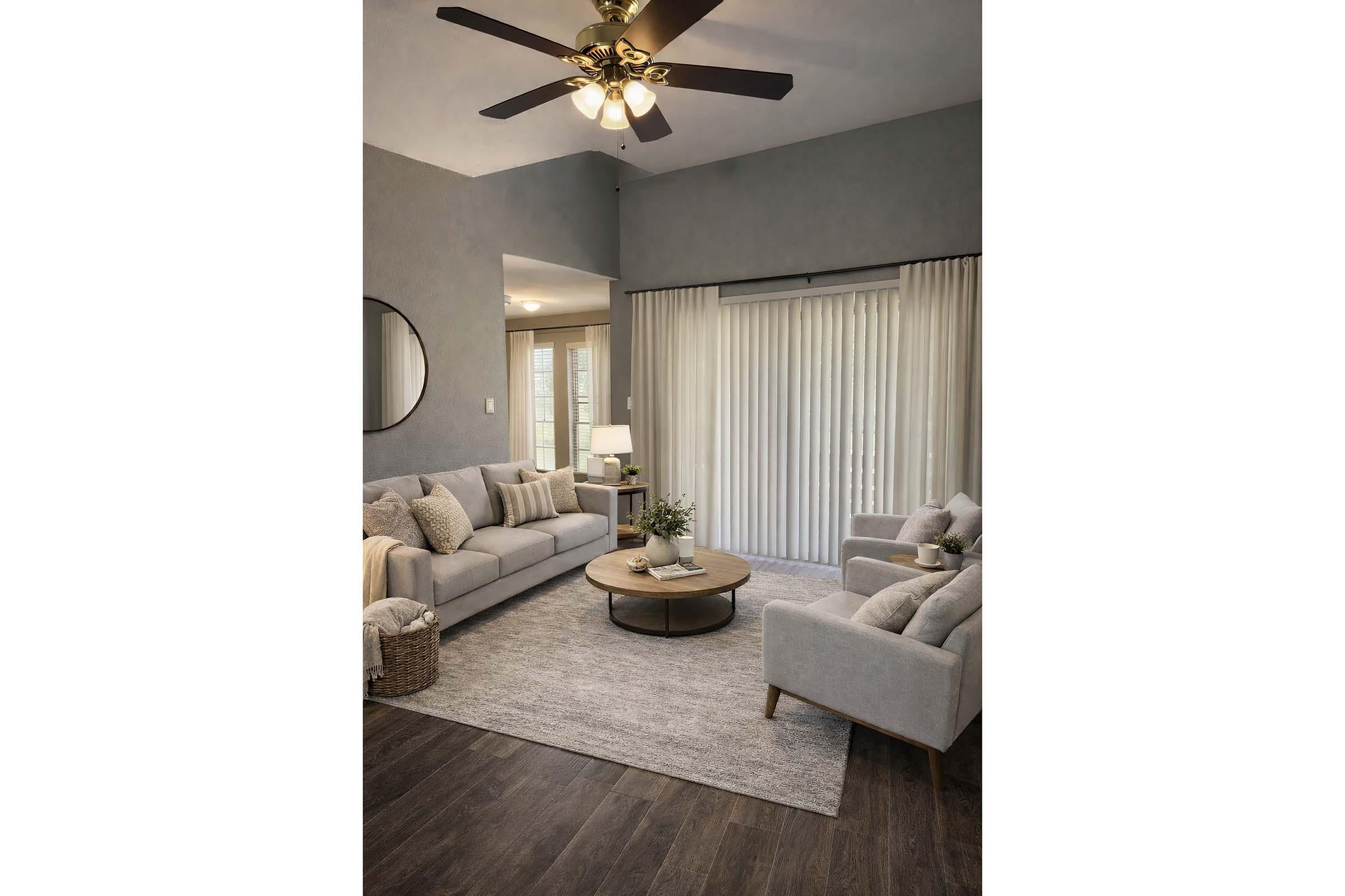 A cozy living room featuring a light gray sofa with decorative pillows, a round coffee table, and two matching armchairs. The space has a neutral color palette, a ceiling fan with lights, and large windows covered by sheer curtains, allowing natural light to fill the room. A stylish rug complements the decor.