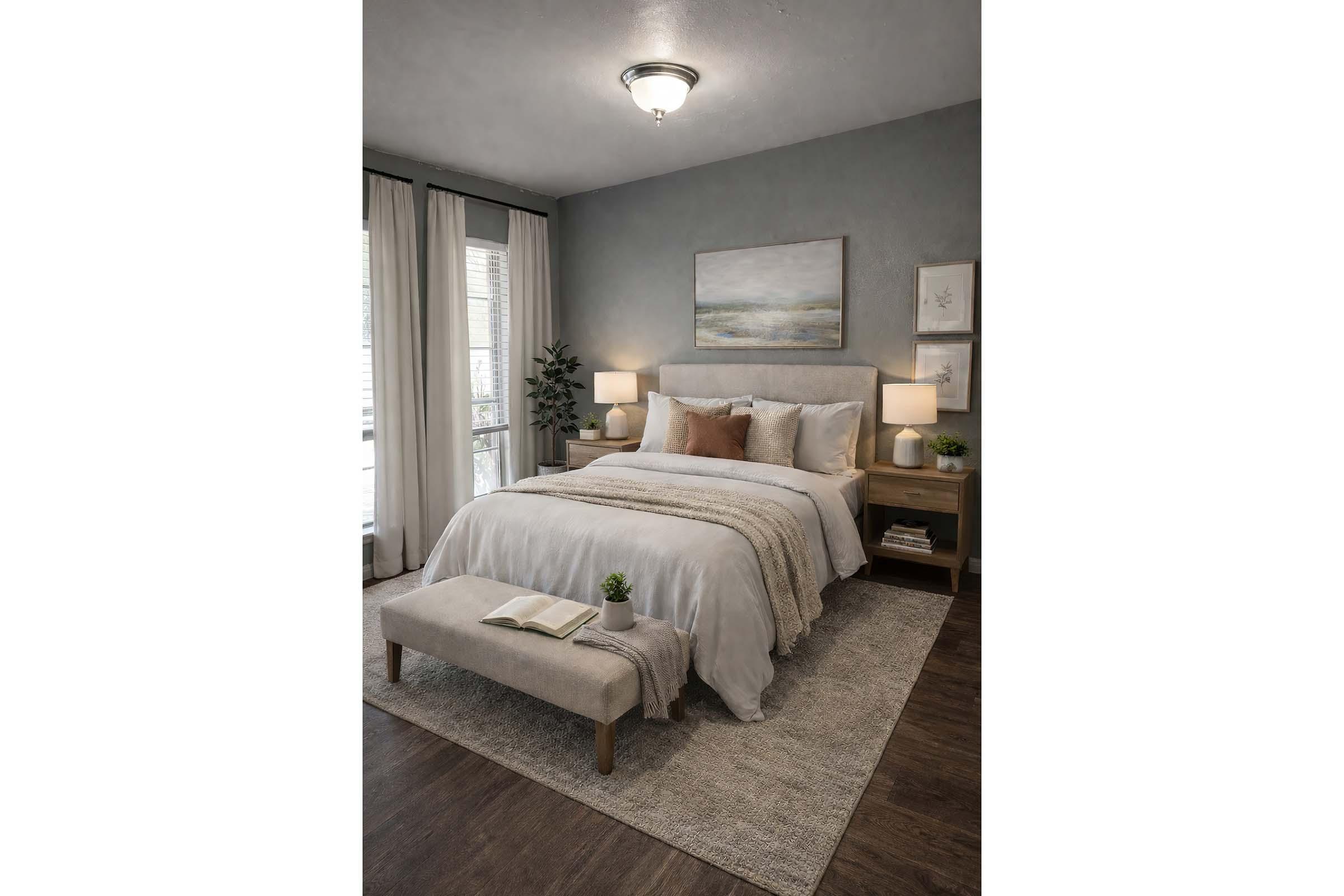 A cozy bedroom featuring a light grey bedspread with accent pillows, a wooden headboard, and bedside tables with lamps. A bench at the foot of the bed and a small plant add warmth. Natural light filters through sheer curtains, and artwork is displayed on the walls, creating a serene and inviting atmosphere.