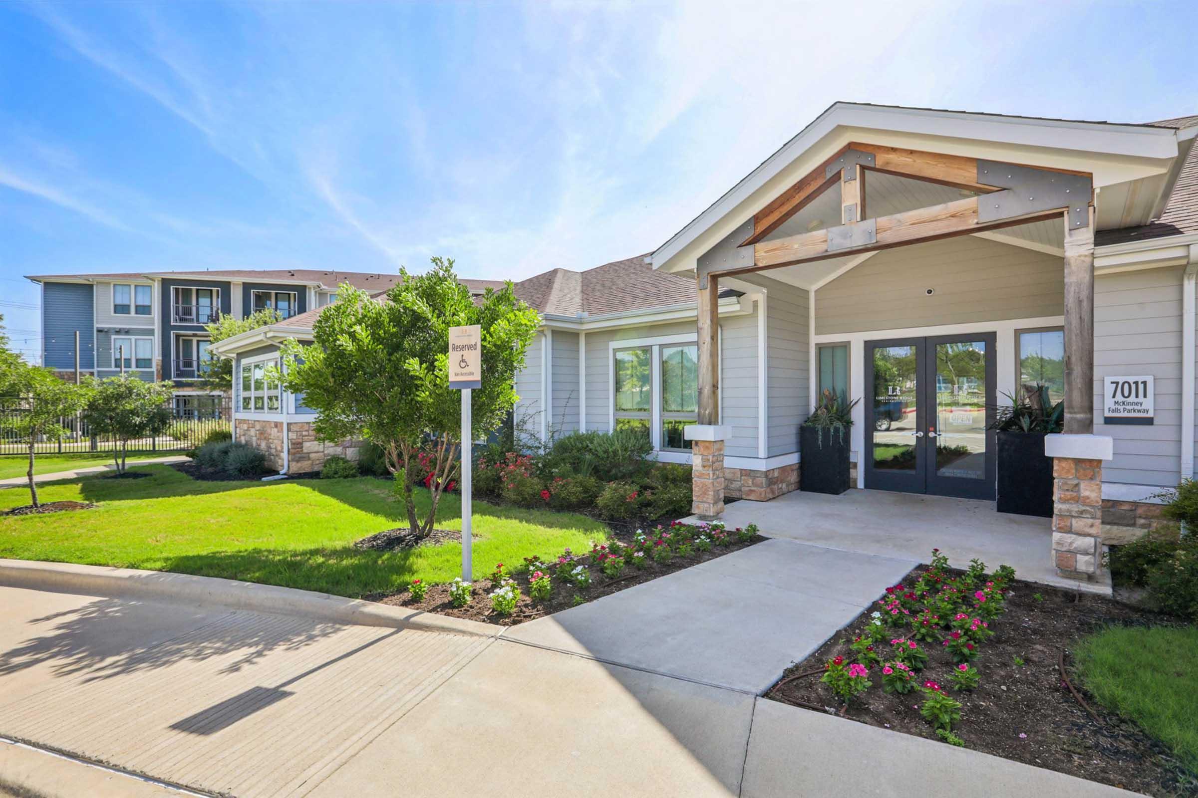 A modern building entrance with a welcoming design featuring large windows and a wooden overhang. Well-maintained landscaping includes green grass, shrubs, and flower beds. A sign is visible near the entrance. In the background, additional residential buildings can be seen.