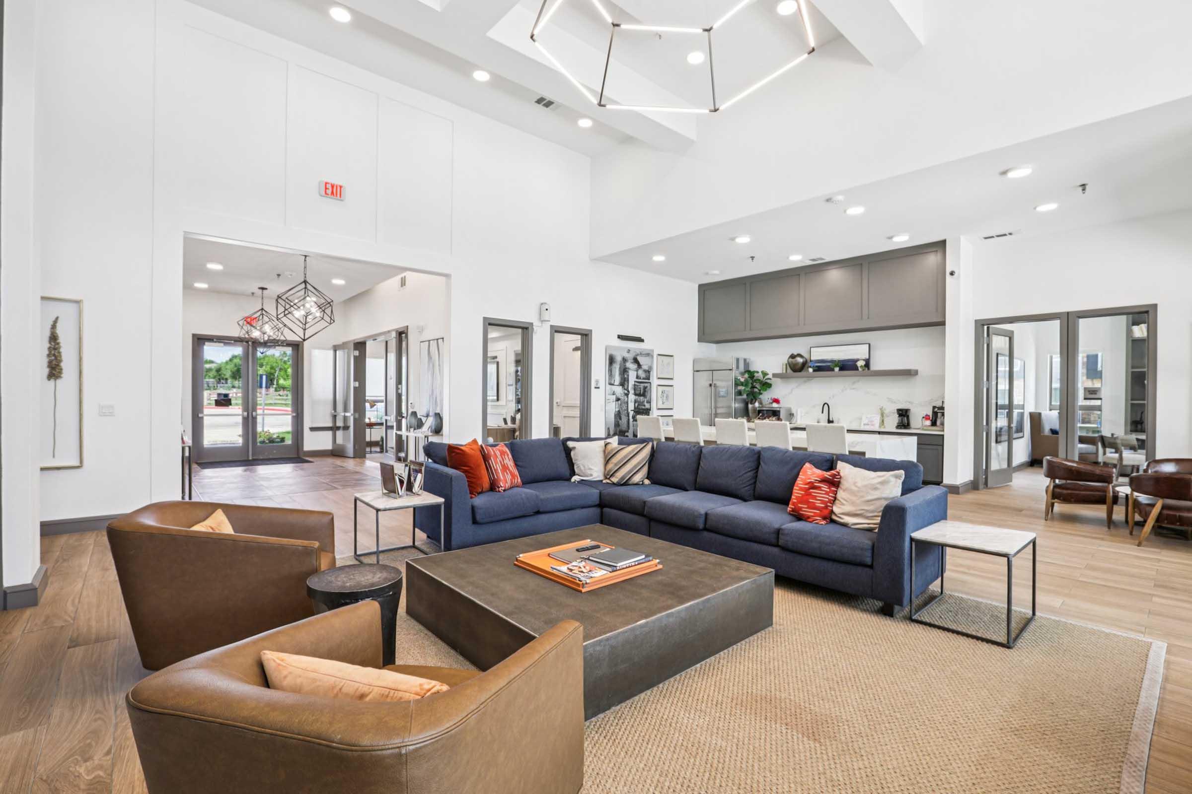Modern interior of a spacious living area featuring a large navy blue sectional sofa with decorative pillows, a dark coffee table, and stylish brown armchairs. The room has high ceilings, ample natural light, and a kitchen visible in the background, with contemporary decor and furnishings.