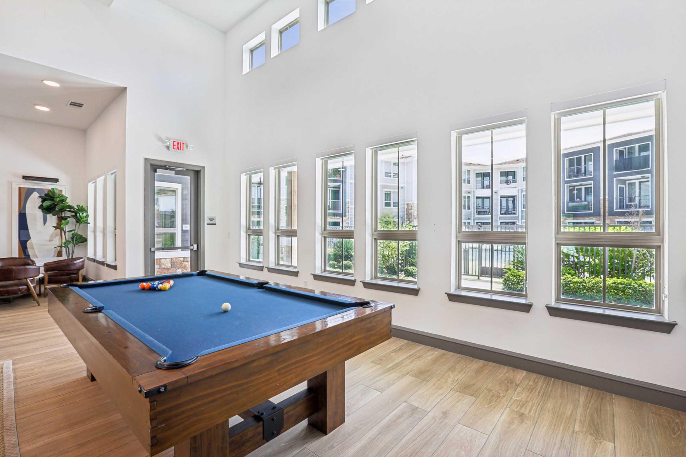 A bright indoor space featuring a billiard table with colored balls, large windows allowing natural light, and green views outside. The room has a modern aesthetic with wood flooring and a few comfortable seating areas.
