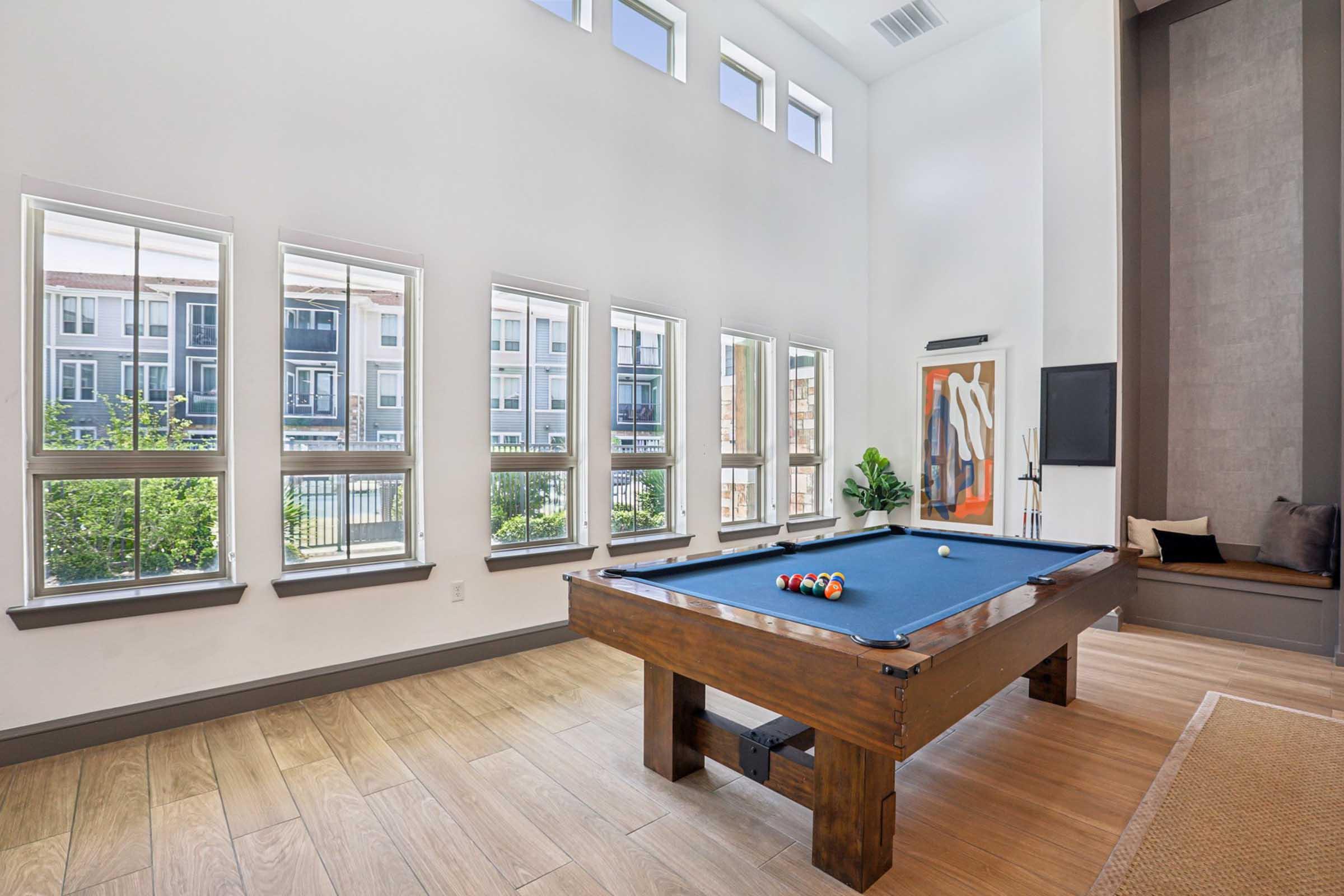 A bright indoor space featuring a billiards table, surrounded by large windows that provide ample natural light. The room has modern decor with a plant and a television mounted on the wall, along with a cozy sitting area in the corner.