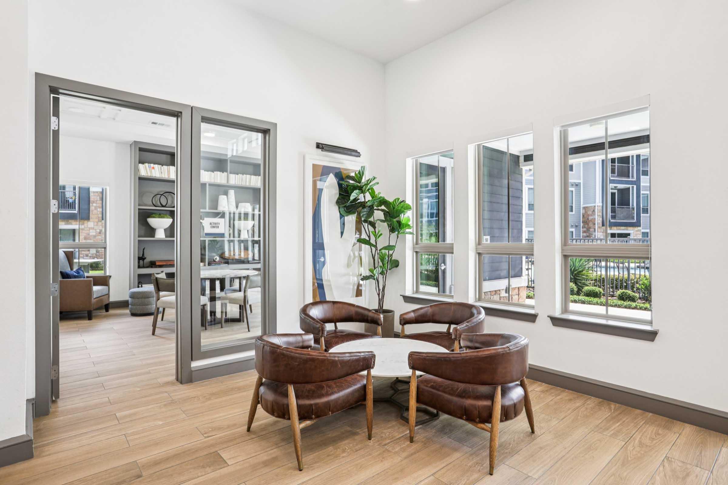 A modern lounge area featuring brown leather chairs surrounding a round table, large windows providing natural light, and a view of greenery outside. A doorway leads to an adjacent room with bookshelves and seating, enhancing the inviting atmosphere. The space has warm wooden flooring and contemporary decor.