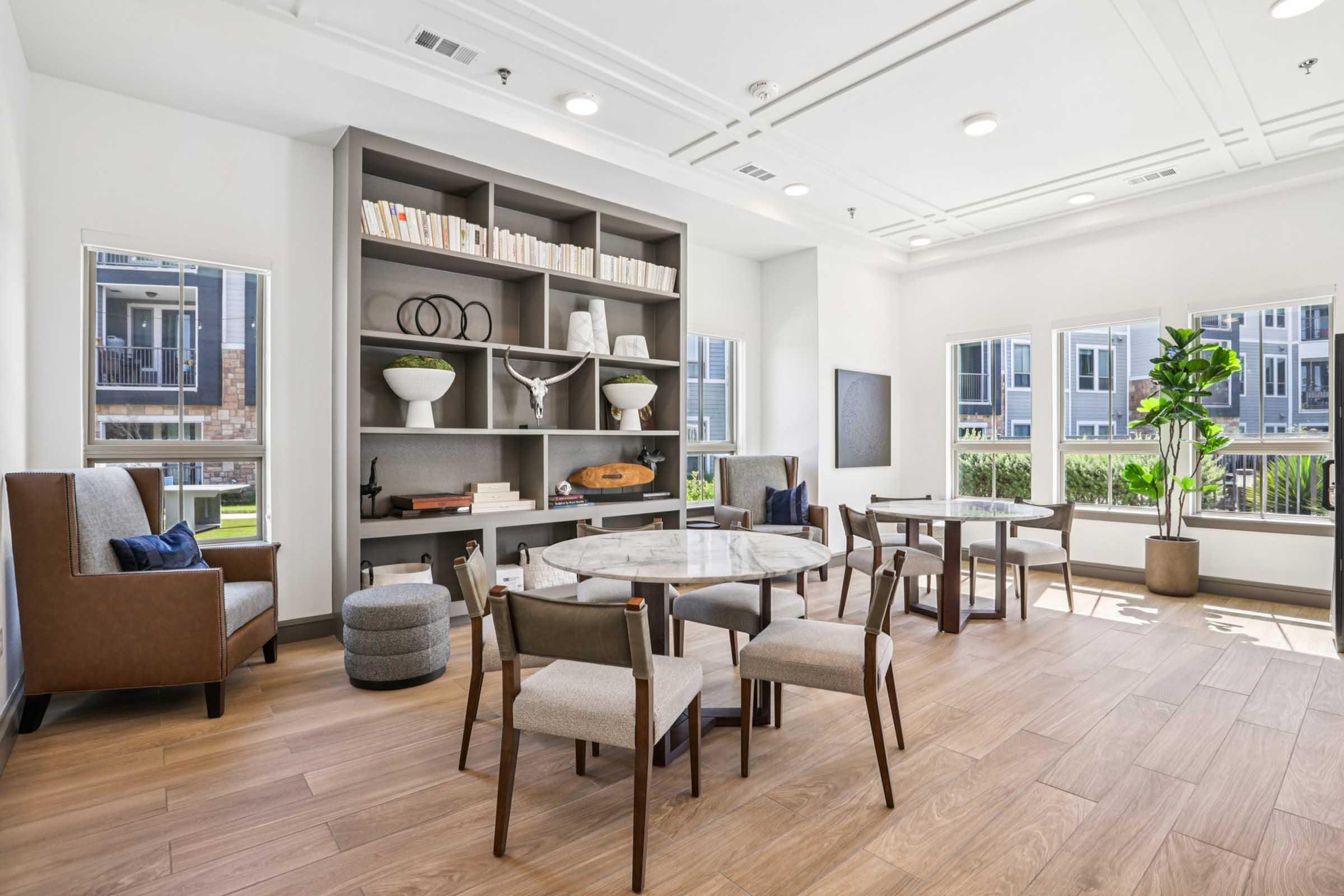 A modern, cozy lounge area featuring a large oval table surrounded by chairs, a built-in bookshelf with decorative items, and large windows allowing natural light. A comfortable seating arrangement includes an armchair and a plant, creating a welcoming atmosphere.