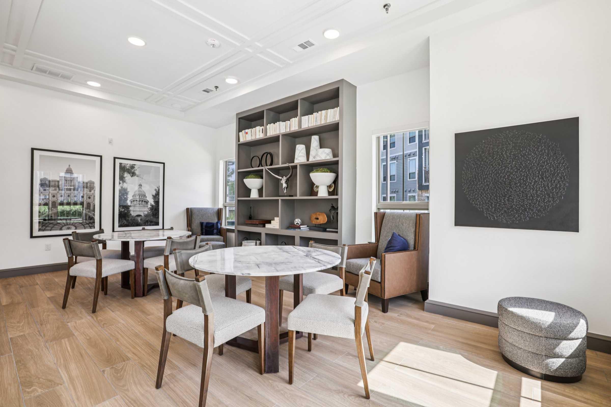 A modern common area featuring a round marble table with four chairs, a bookcase filled with books and decorative items, and an abstract wall art piece. Large windows let in natural light, enhancing the warm wood flooring and contemporary furnishings.