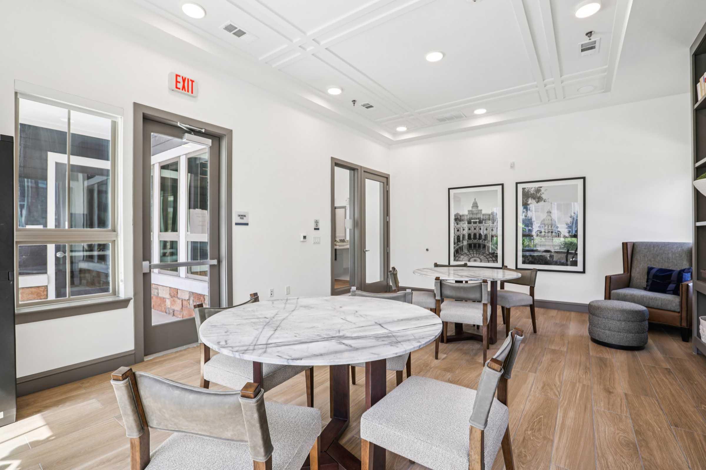 A modern interior room featuring a marble-top round table surrounded by four upholstered chairs. There are two framed photographs on the wall depicting architectural landmarks. A door leads outside, and a cozy seating area with an armchair is visible in the corner. The decor is bright and contemporary.