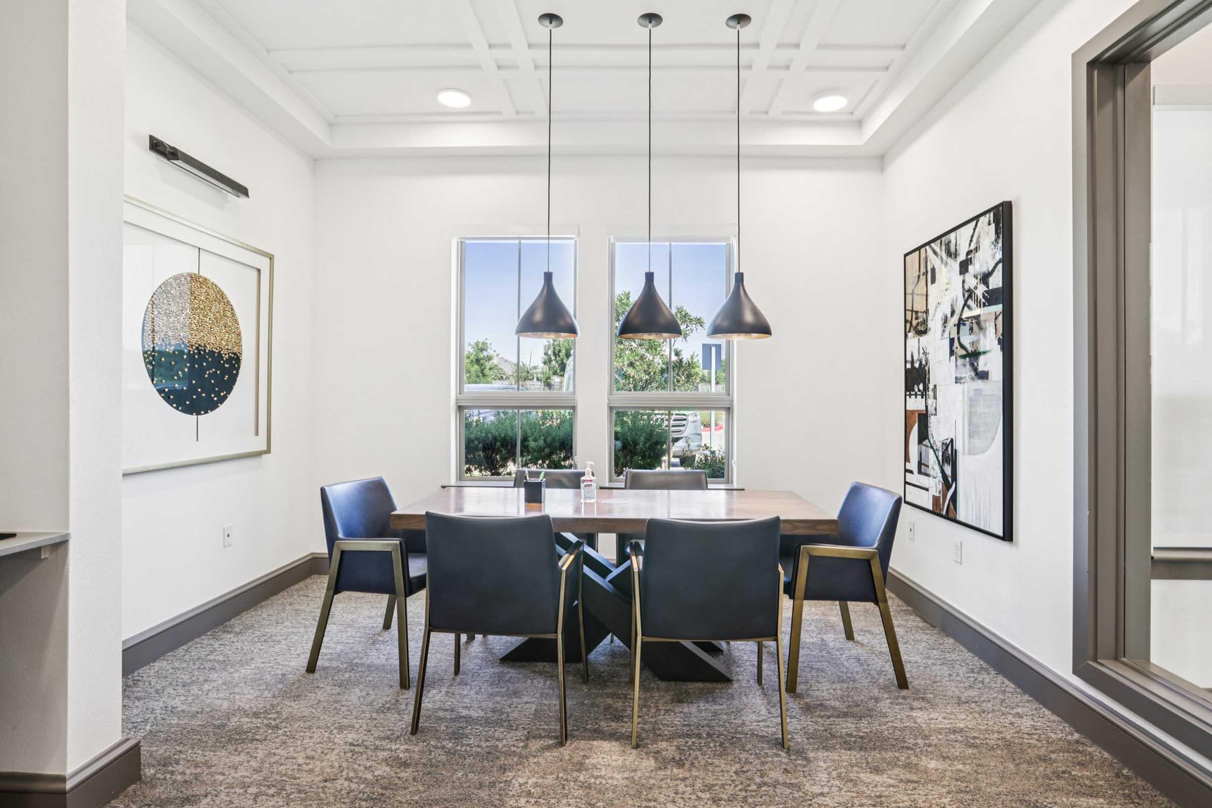 A modern meeting room featuring a large wooden table surrounded by four blue chairs. Two framed artworks decorate the walls, and three pendant lights hang from the ceiling, providing bright illumination. Large windows let in natural light, creating a welcoming atmosphere.