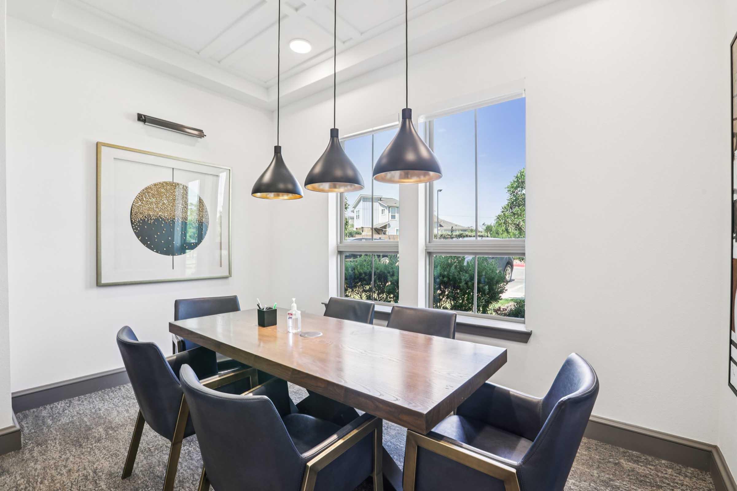 A modern conference room featuring a wooden table surrounded by dark blue chairs. Three pendant lights hang above the table, and a large framed artwork with a golden circular design is on the wall. Large windows allow natural light to flood the room, with greenery visible outside.