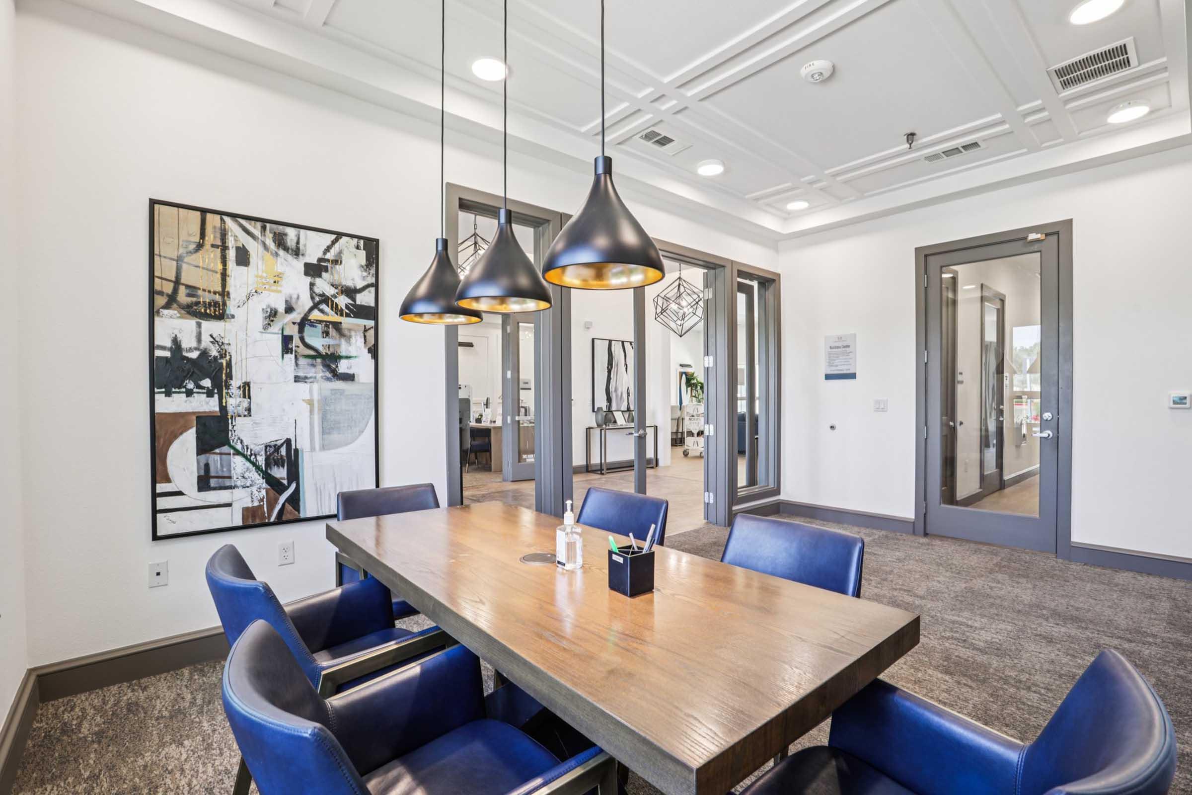 A modern office meeting room featuring a large wooden table surrounded by blue leather chairs. Three pendant lights hang above the table. The room has a contemporary art piece on the wall and large windows that lead to an adjoining workspace. The overall design is sleek and professional.