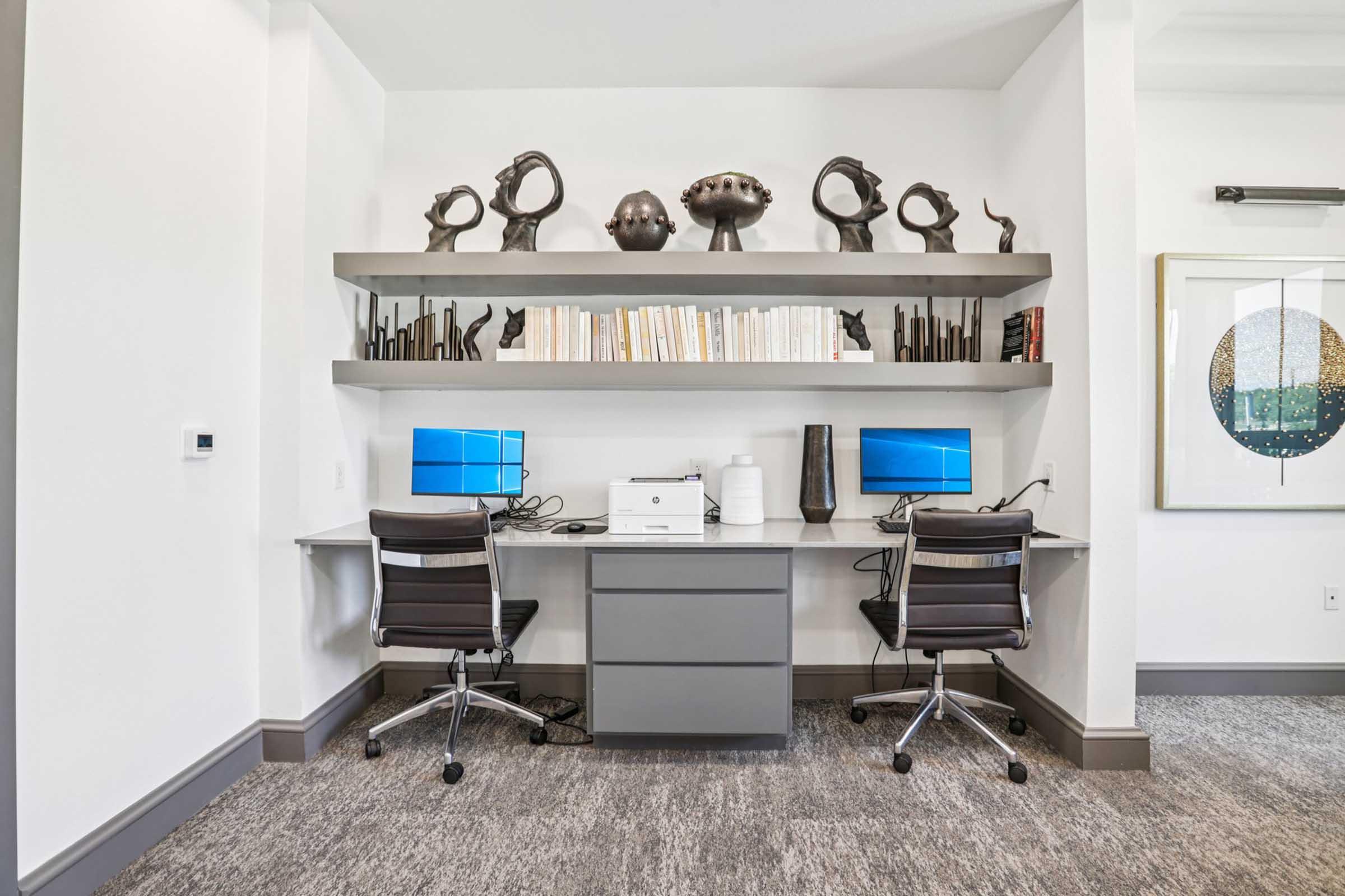 A modern workspace featuring a sleek desk with two computer monitors and a printer. Above, there are decorative sculptures on a shelf, alongside neatly arranged books. The room has a clean, minimalist design with white walls and a contemporary feel.