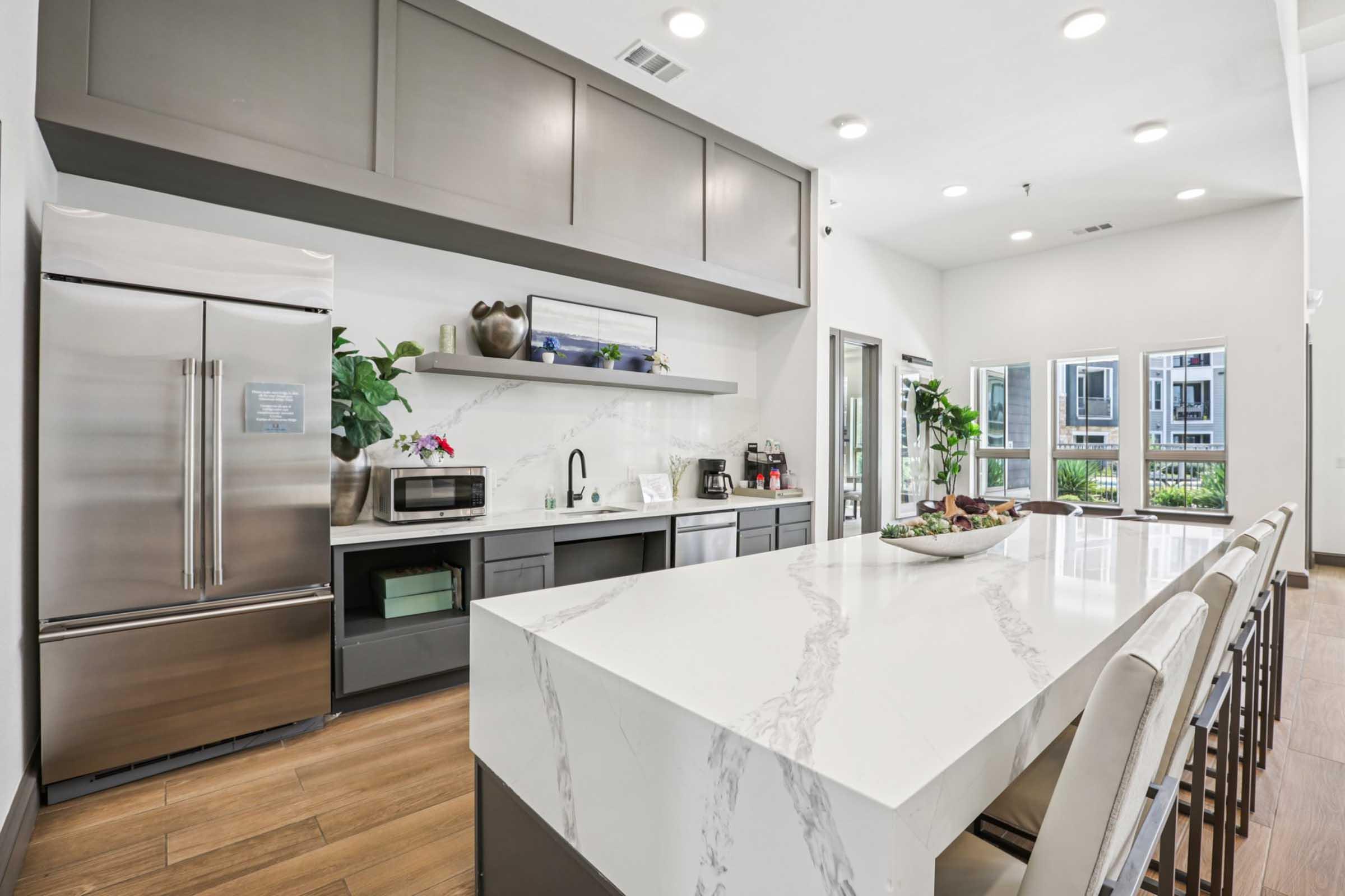 Modern kitchen featuring a large island with a marble countertop and bar stools. Stainless steel appliances, including a refrigerator and microwave, are complemented by stylish cabinetry. The space is brightened by large windows, showcasing greenery outside. Elegant decor adds a contemporary touch.