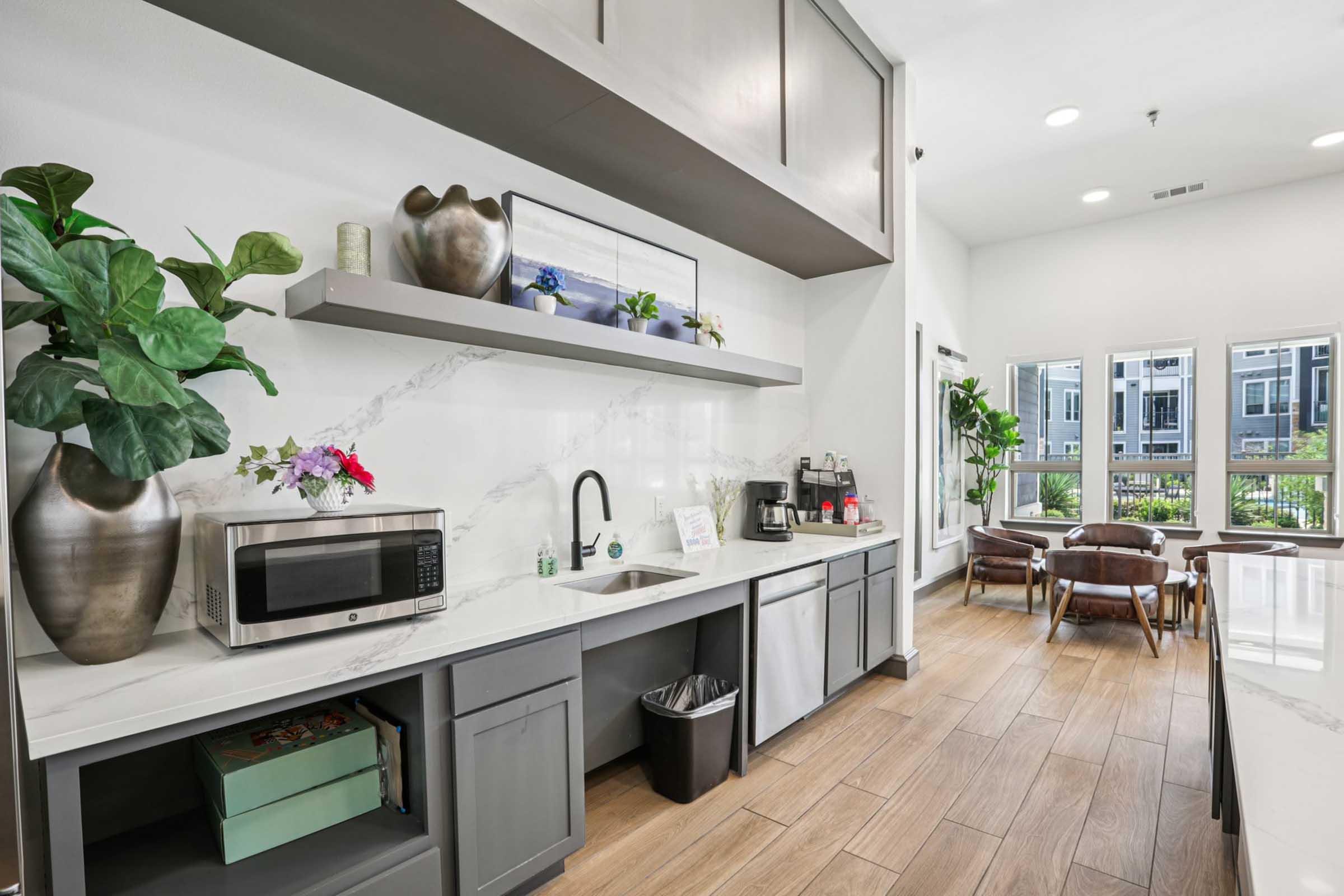 A modern kitchen area featuring gray cabinetry, a stainless steel sink, a microwave, and decorative plants. There are shelves adorned with decorative items and flowers. Large windows allow natural light, revealing a view of an outdoor area with greenery and furniture.