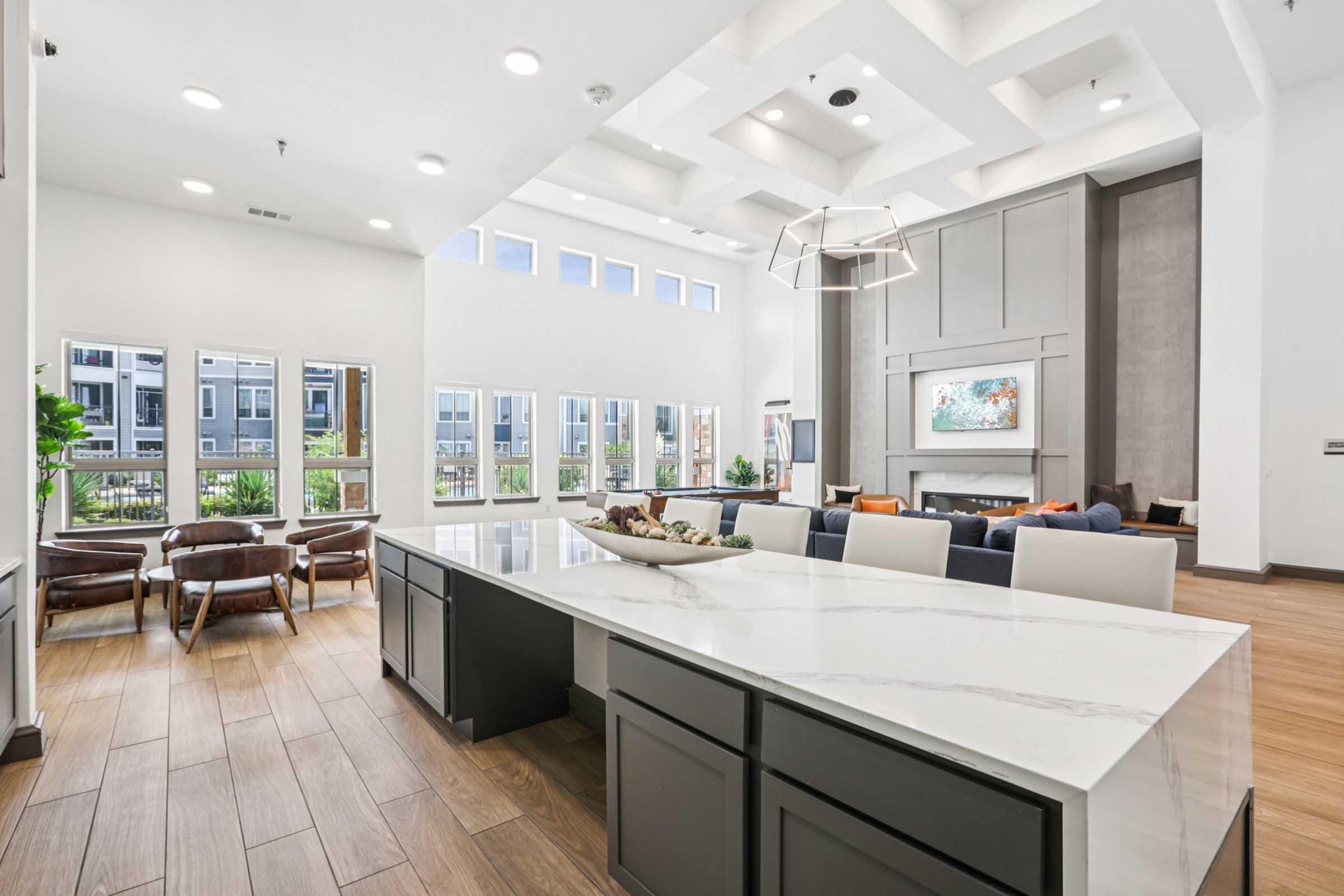 A bright, modern interior view of a spacious living area featuring a large white kitchen island with a decorative bowl, comfortable seating, large windows allowing natural light, and stylish decor. The room has a contemporary design with warm wood flooring and a cozy fireplace.