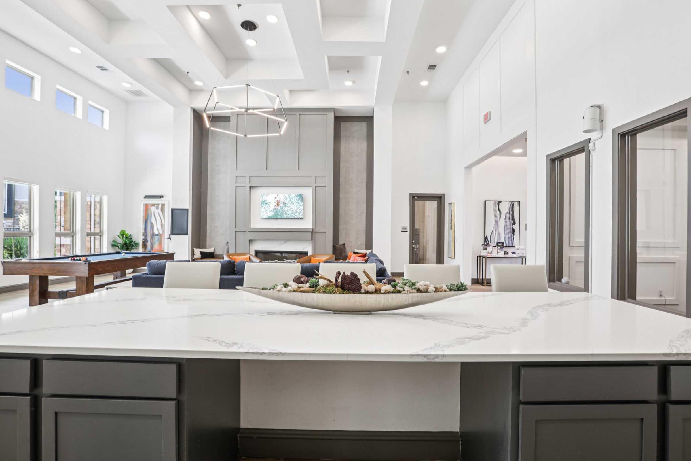 A modern living space featuring a large open layout with a stylish kitchen island, marble countertop, and minimalist decor. The room includes a cozy seating area, a television, and large windows allowing natural light to fill the space. There are decorative elements displayed on the island.