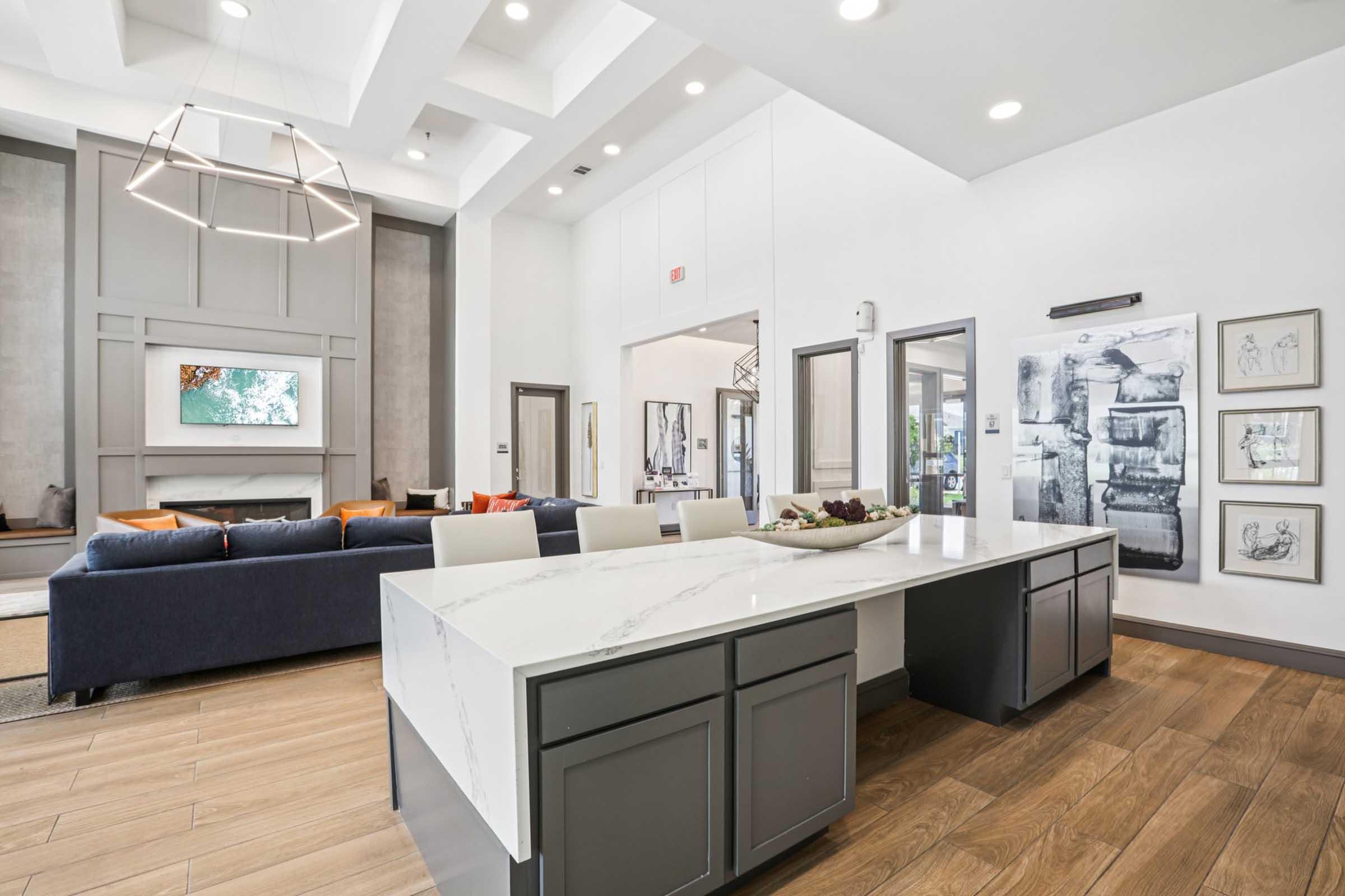 Modern kitchen and dining area featuring a large marble island with dark cabinetry, a spacious living room in the background with a blue sofa and colorful cushions, and wall decor including framed artwork. The room is bright with natural light and high ceilings.