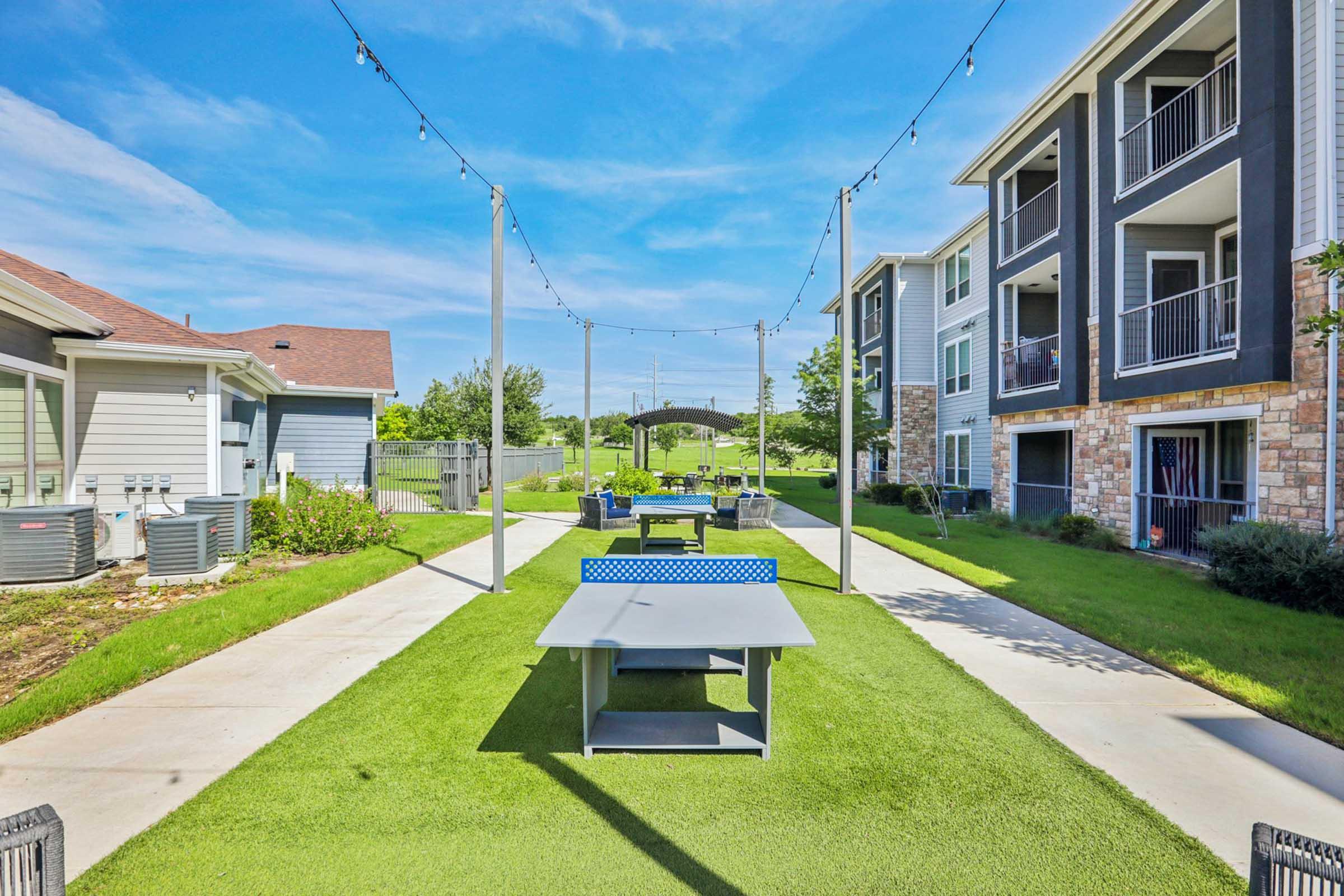 A well-maintained outdoor area featuring several picnic tables on green grass, surrounded by trees and apartment buildings. Warm sunlight illuminates the space, with string lights overhead adding a charming touch. Ideal for socializing and relaxation.