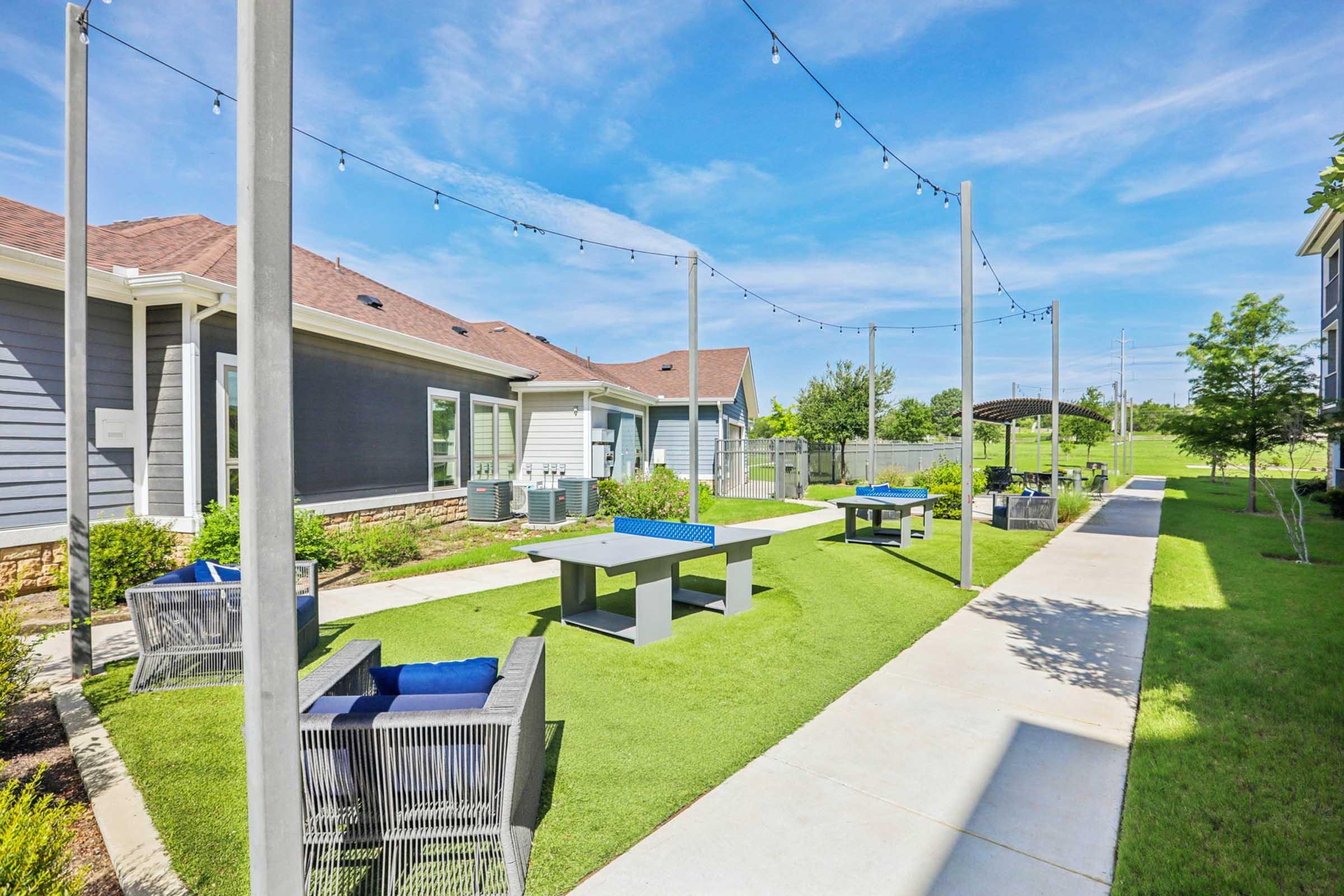 A well-maintained outdoor area featuring green lawns, paved pathways, and seating arrangements. The space includes blue tables and chairs under string lights, with modern buildings in the background. Clear blue skies give the setting a bright and inviting atmosphere.