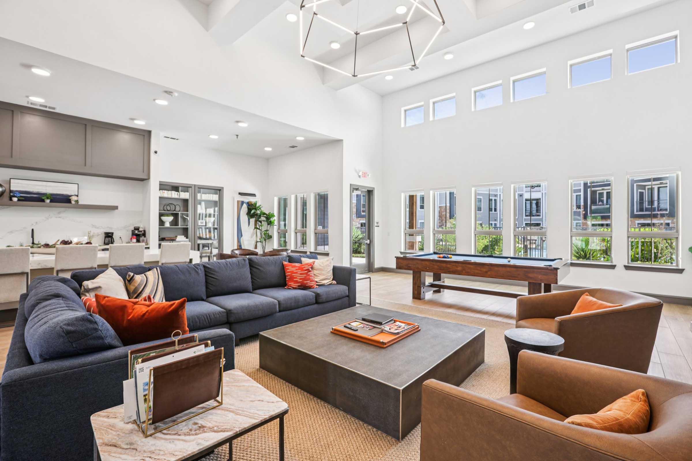 A modern, spacious living area with large windows allowing natural light. Features a comfortable sectional sofa with colorful cushions, a coffee table in the center, and two stylish armchairs. A sleek kitchen area is visible in the background, along with a pool table near the windows, creating a cozy yet elegant atmosphere.